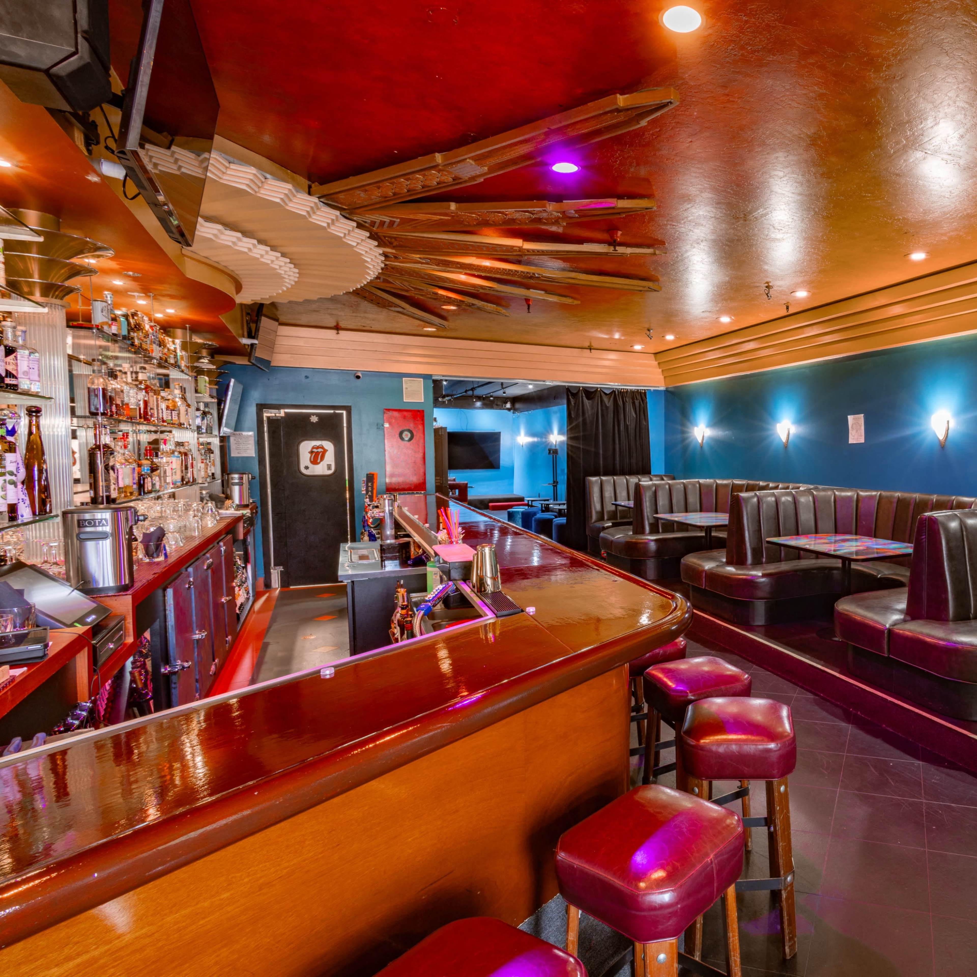 A brightly lit bar area with dark seating booths and a wooden counter, featuring shelves lined with various bottles.