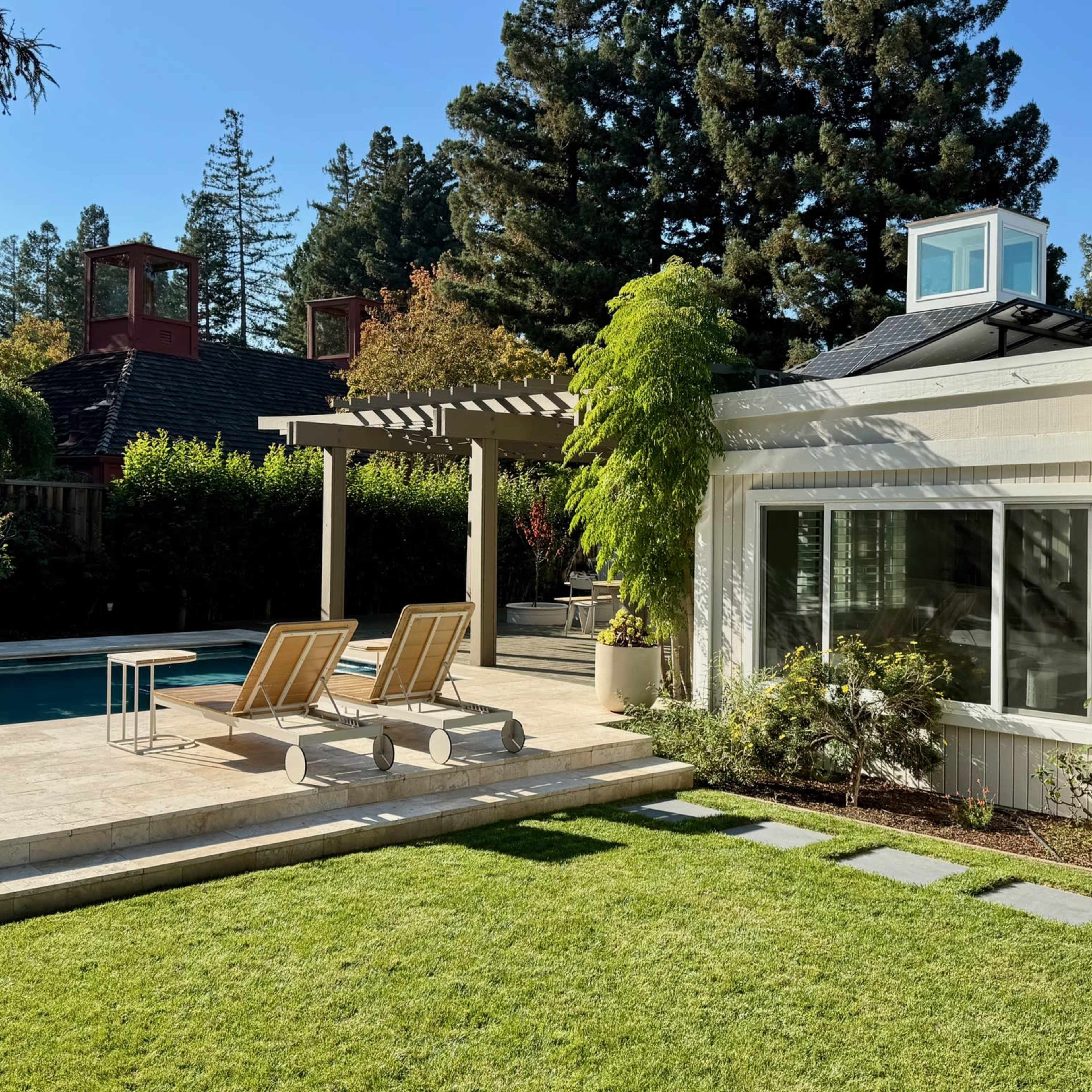 A backyard scene features a swimming pool, two lounge chairs, and a patio area surrounded by greenery and trees.