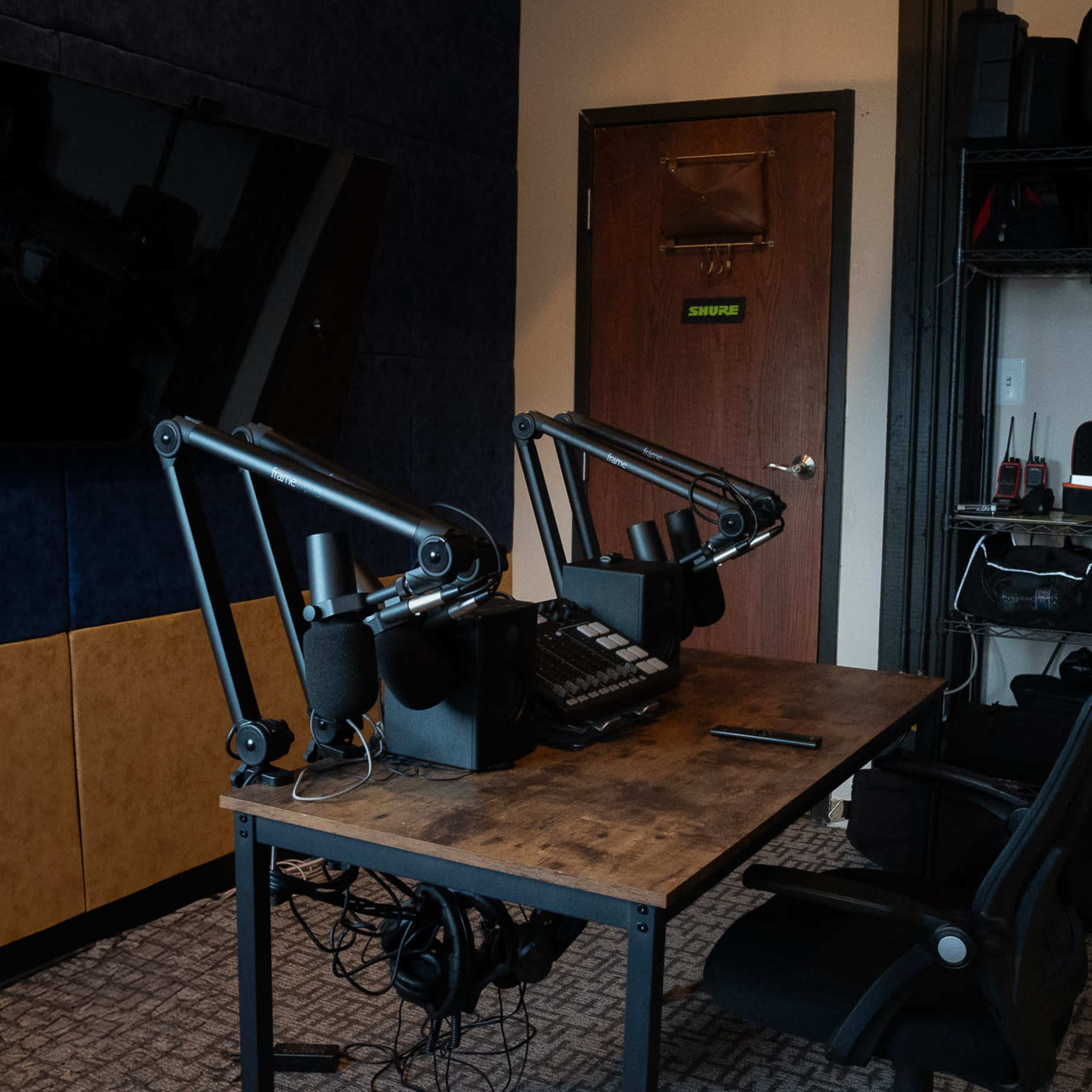 A podcast recording studio features a wooden table with audio equipment, a large television mounted on the wall, and a shelf filled with various gear.