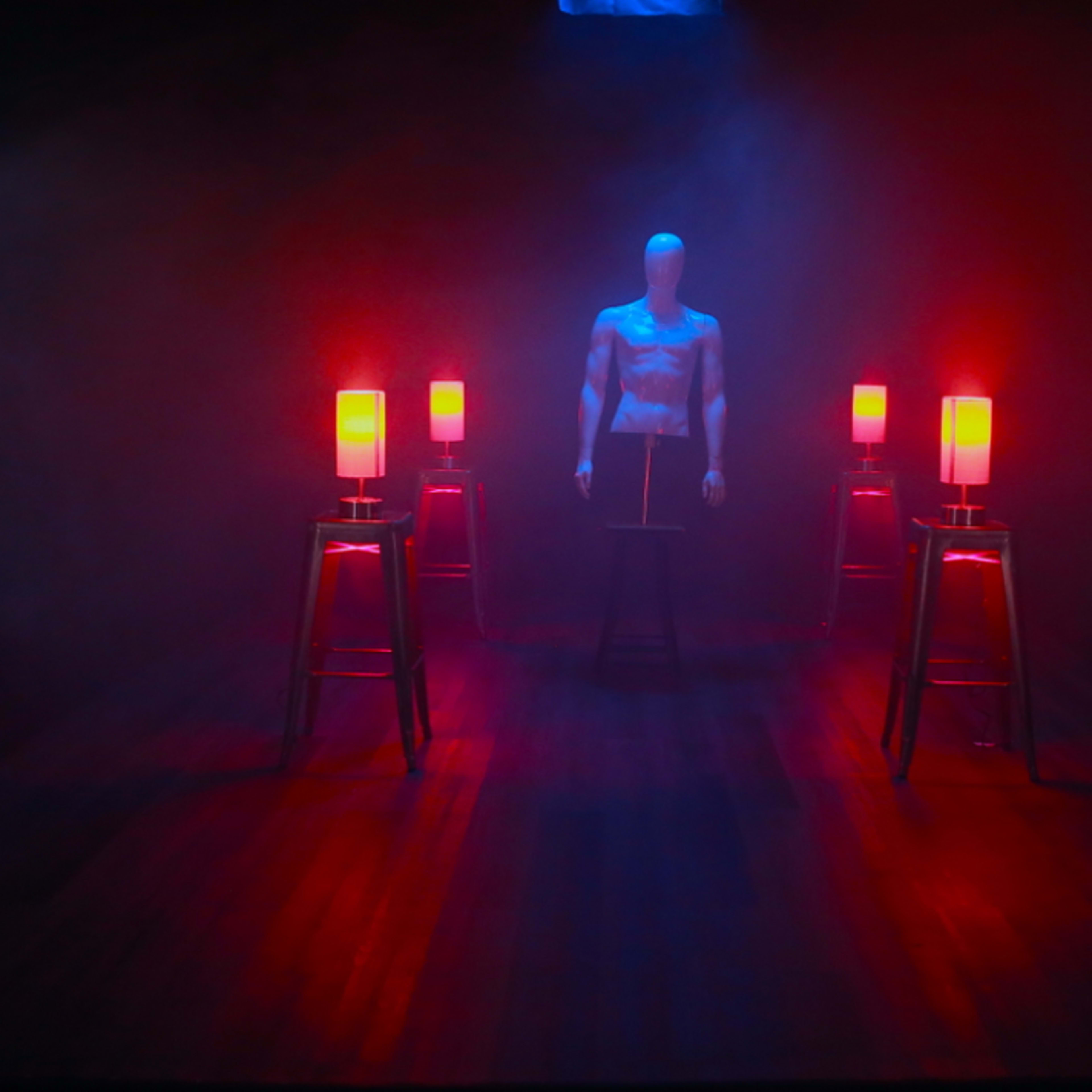 A mannequin stands on a stage surrounded by four illuminated stools, with a foggy atmosphere and red and blue lighting.
