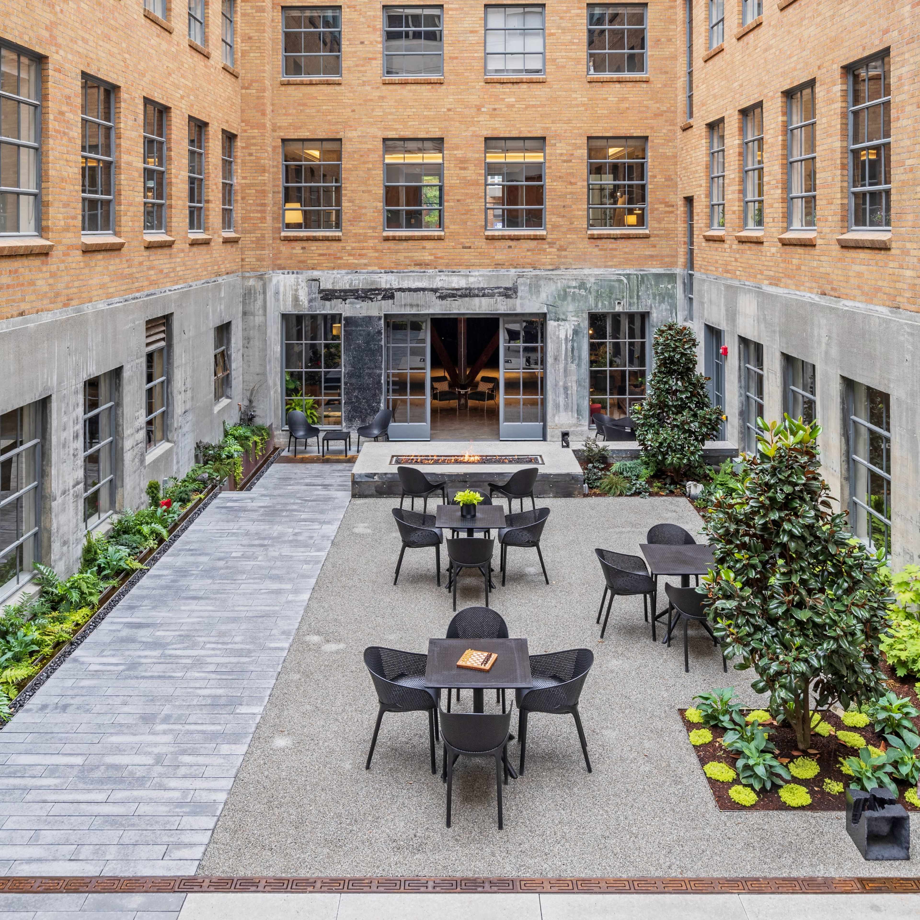 A courtyard with several black tables and chairs, surrounded by greenery and brick buildings.