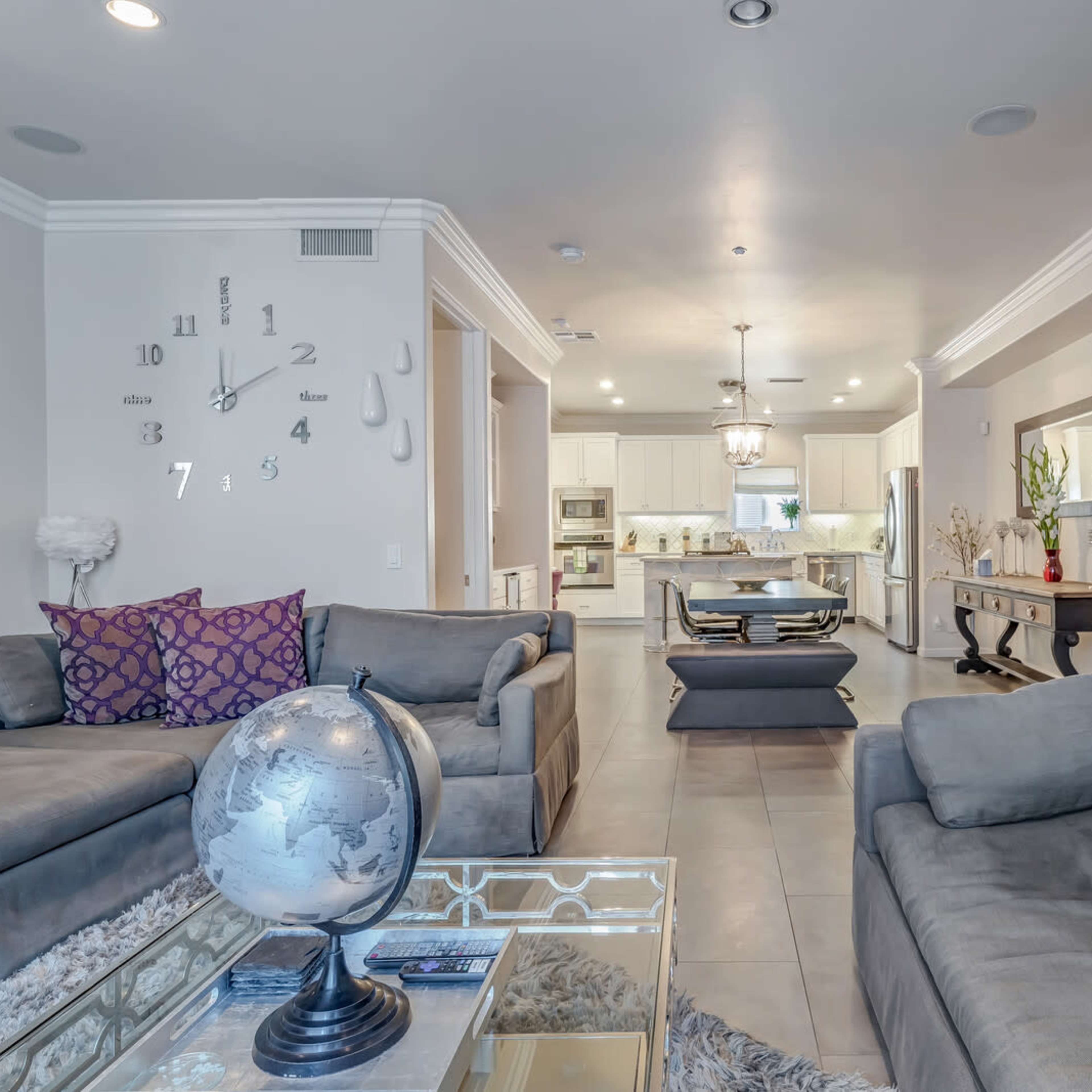 A modern, open-concept living room and kitchen area featuring grey sofas, decorative pillows, a glass coffee table, and a wall clock.