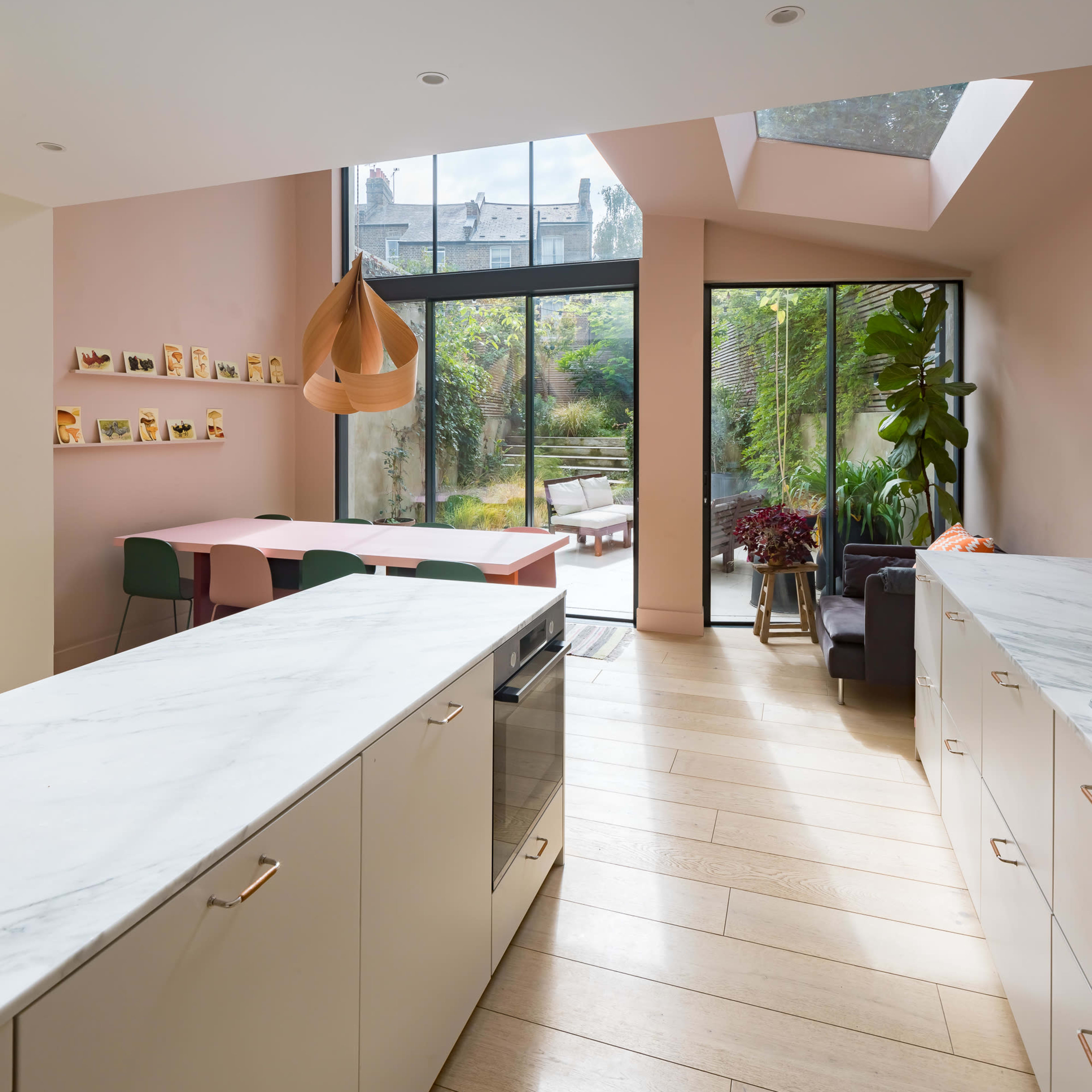A modern kitchen features marble countertops, light-colored cabinetry, and large windows overlooking a garden.
