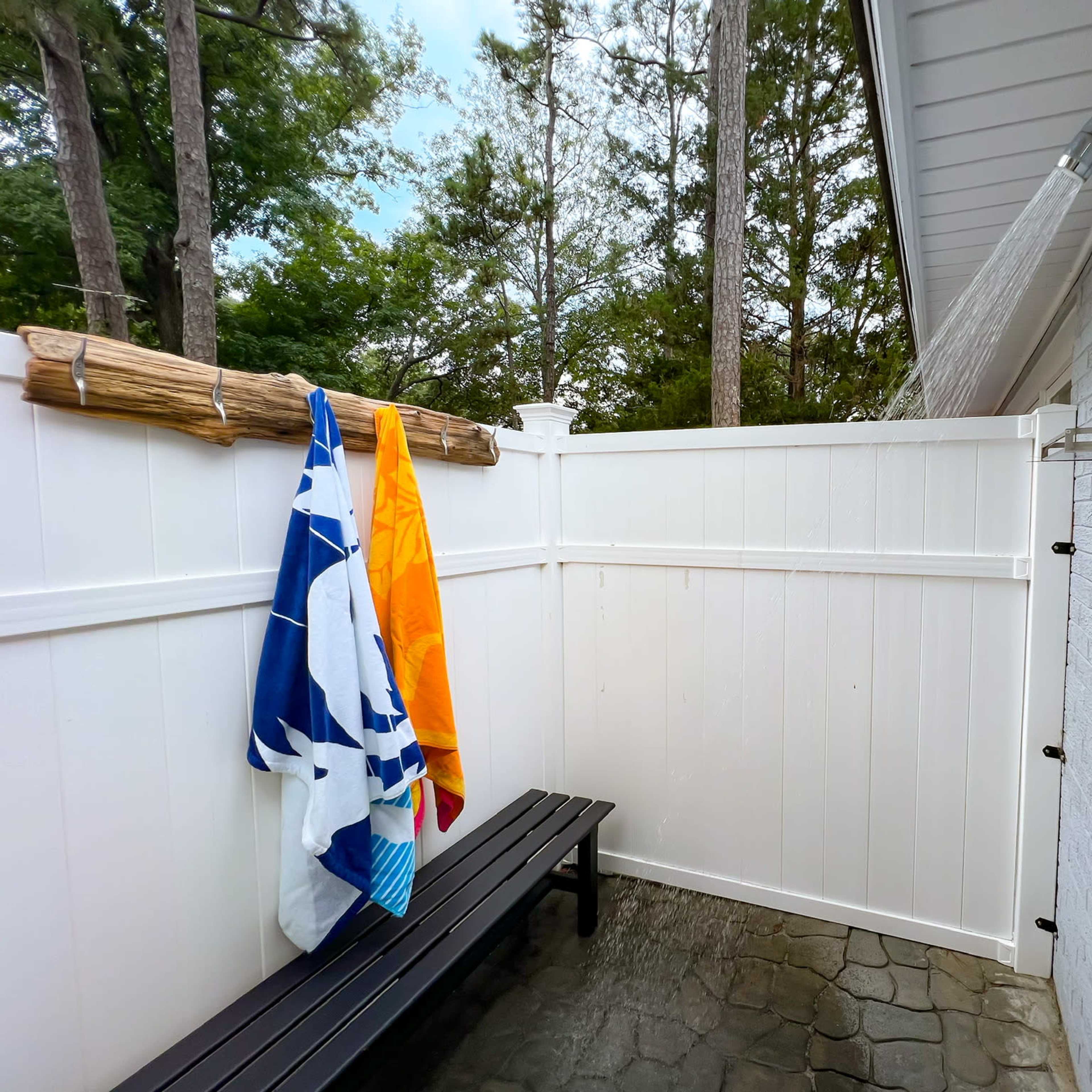 A private outdoor shower area enclosed by white fencing, featuring a bench and two towels hanging on a bamboo rod.