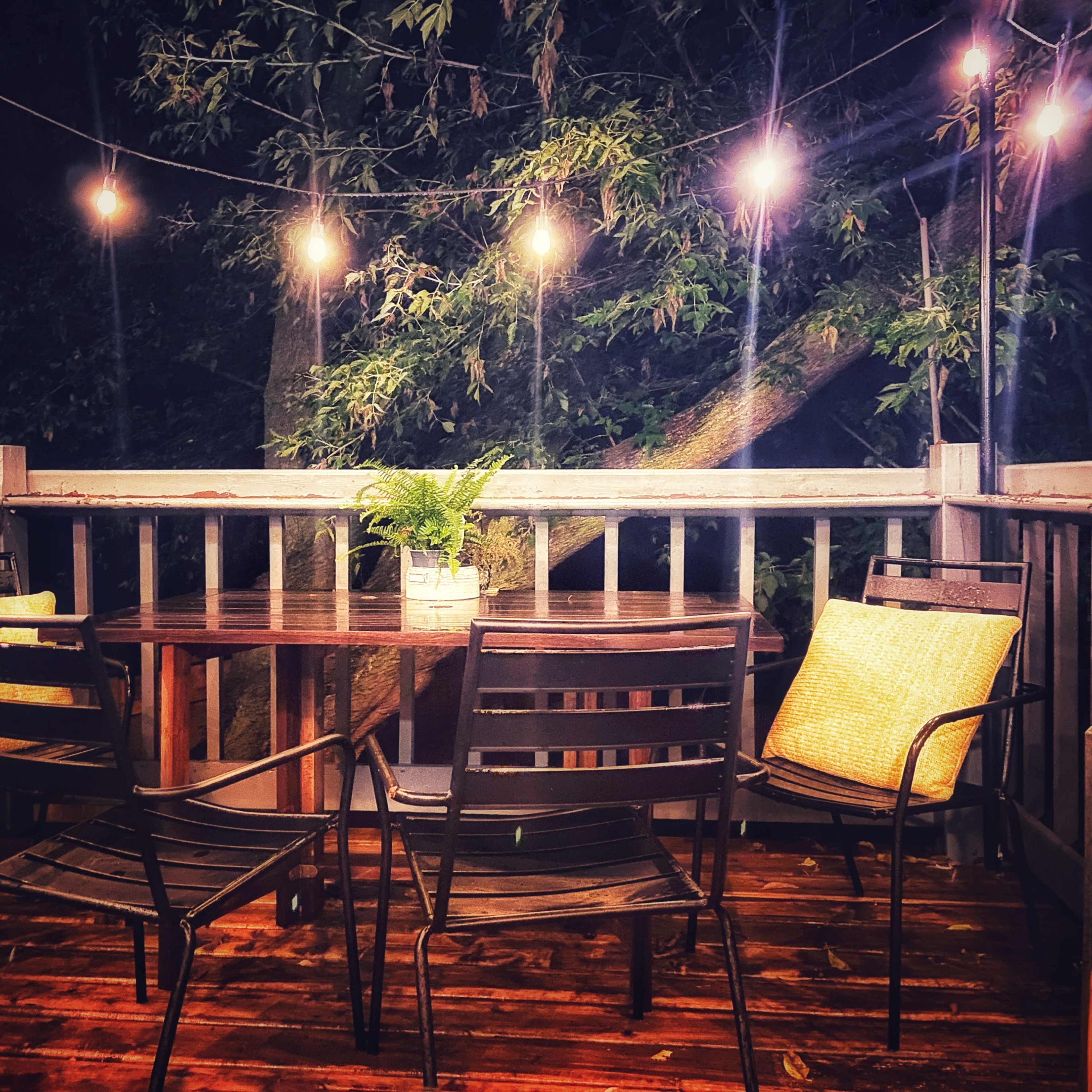 A cozy outdoor deck features a round table surrounded by four black metal chairs, illuminated by string lights overhead.