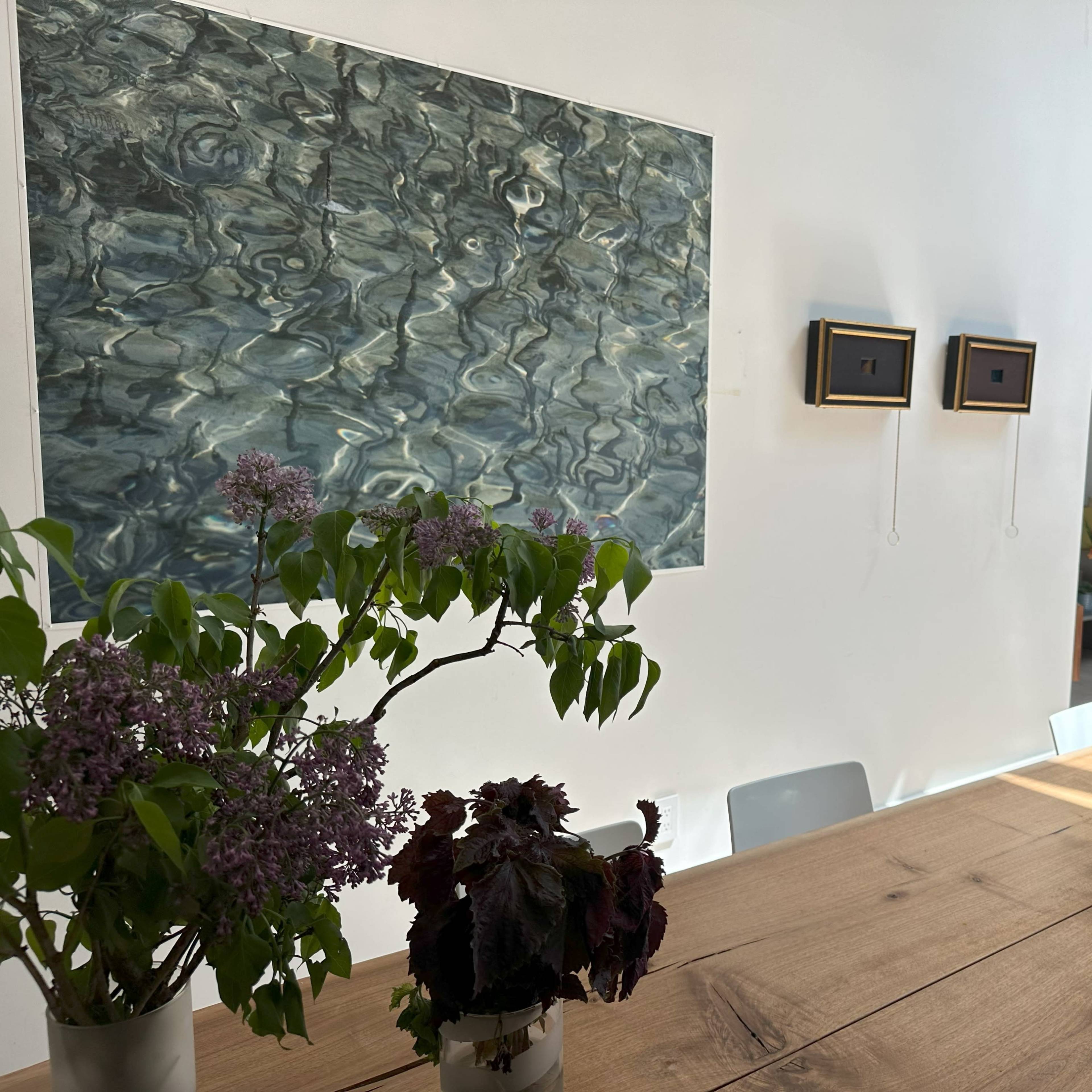 A bright, modern dining area with a wooden table featuring potted plants, a wall art piece depicting water, and a small decorative arrangement on the right.