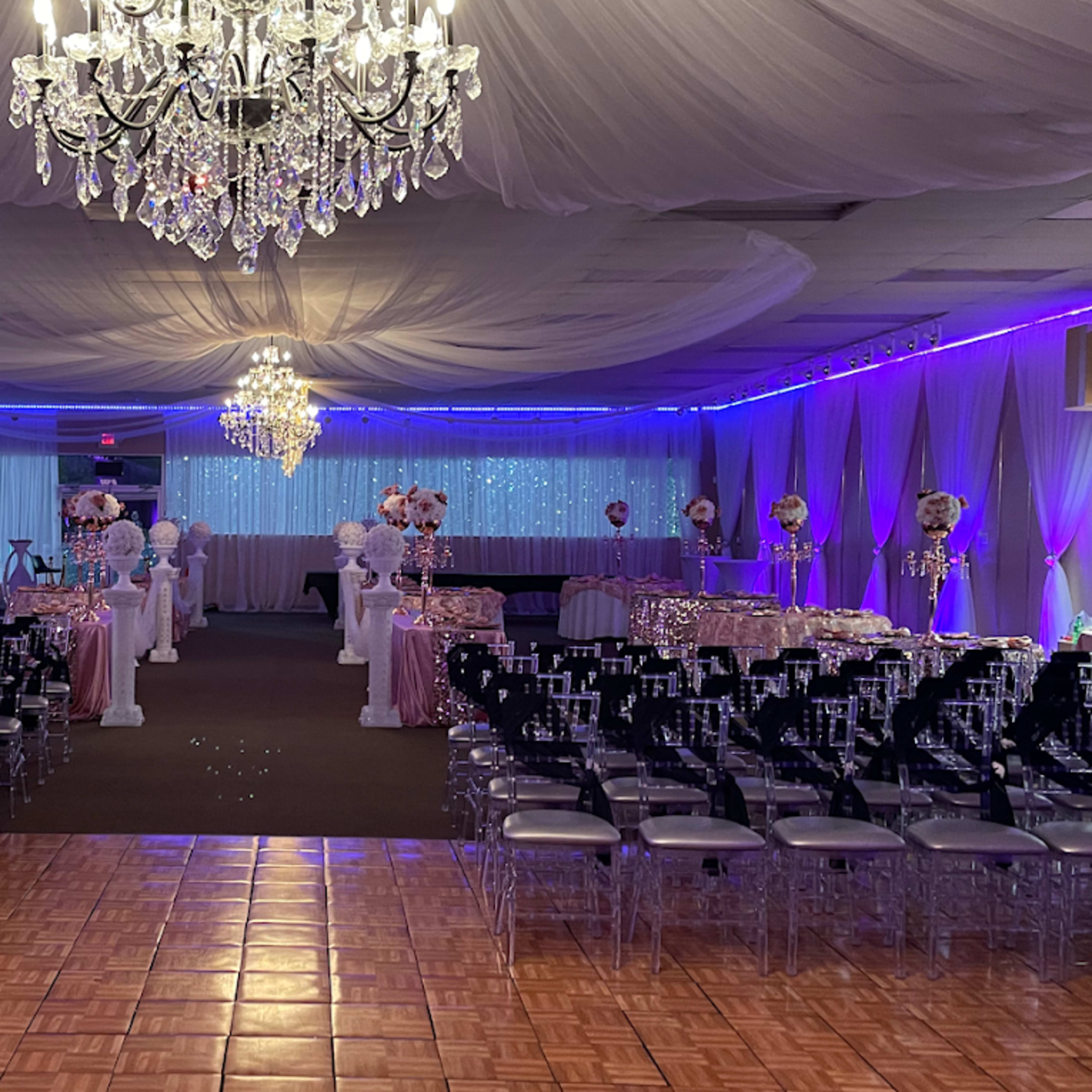 A decorated event venue set up for a formal gathering, featuring rows of chairs, elegant table arrangements, and ceiling drapery with chandeliers.