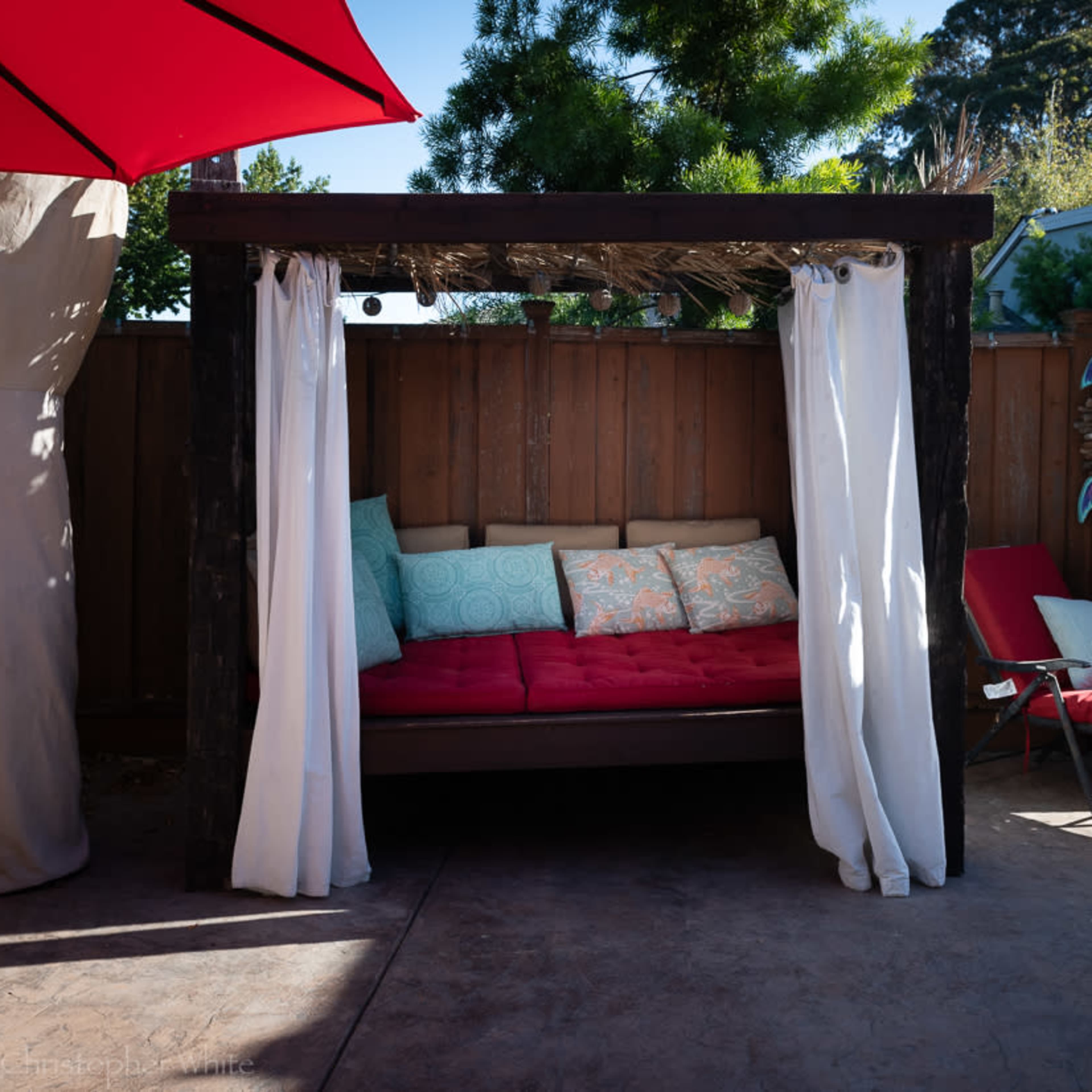 A covered outdoor seating area with colorful cushions, draped curtains, and lounge chairs is set in a backyard surrounded by trees and fencing.