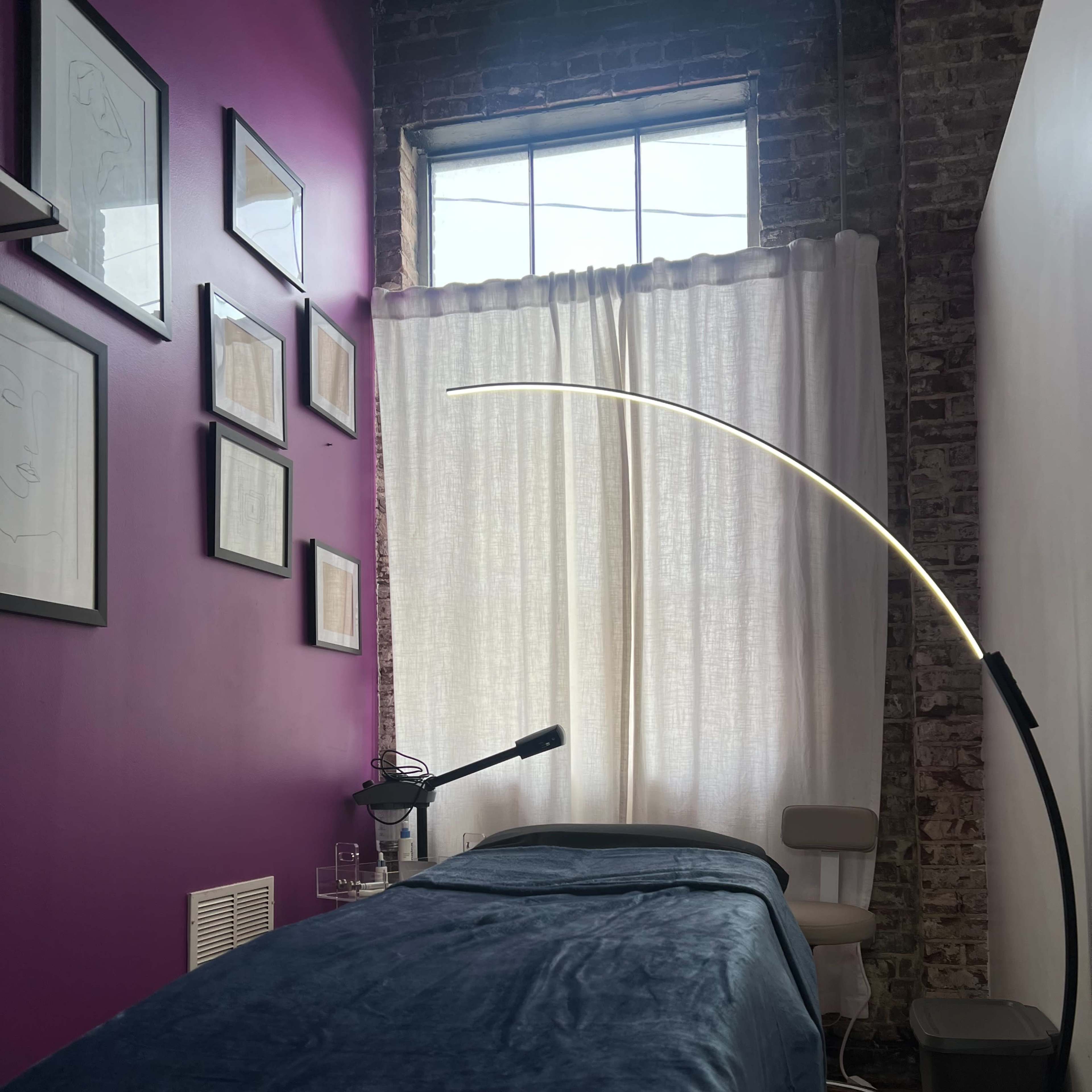 A room with a purple wall featuring framed artwork, a massage table covered with a blue blanket, and a tall lamp beside a chair near a window with sheer curtains.