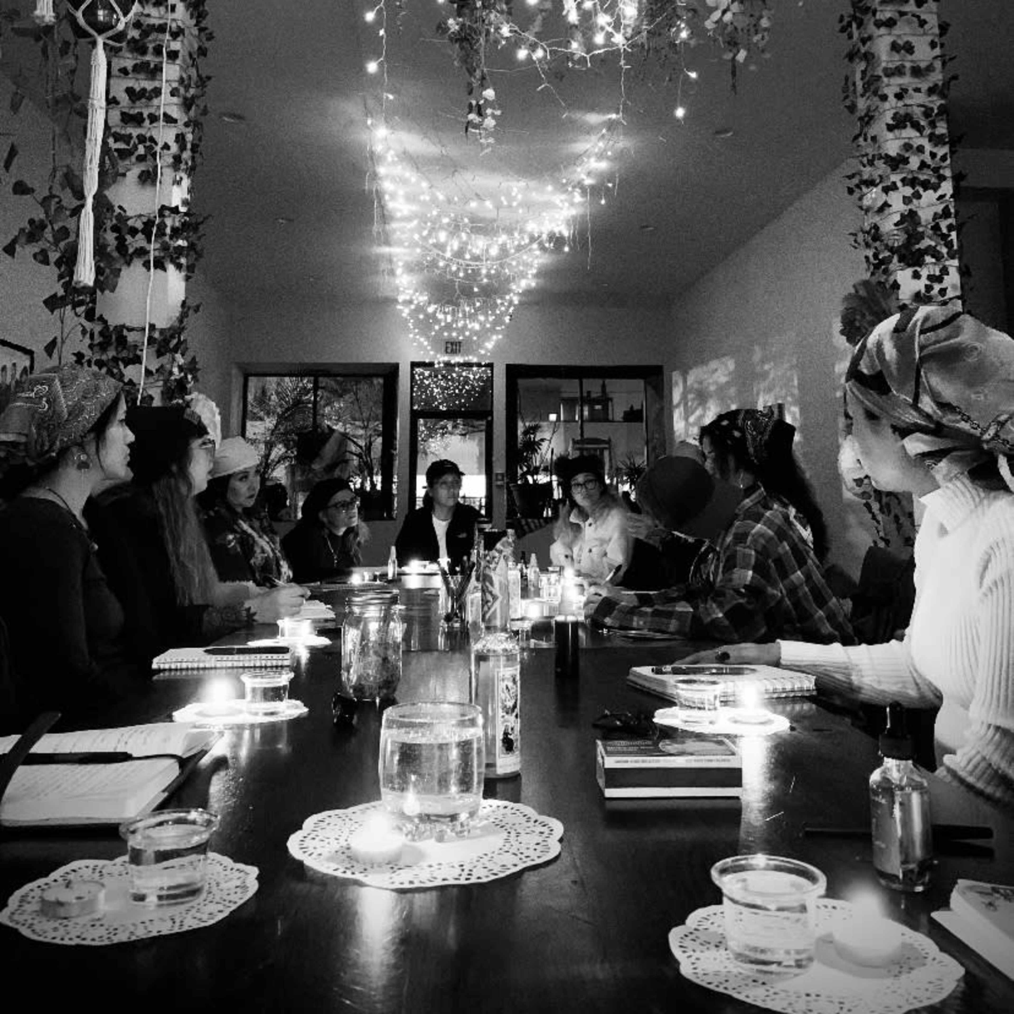 A group of people sits around a long table adorned with candles and foliage, while strings of lights hang above in a warmly lit room.