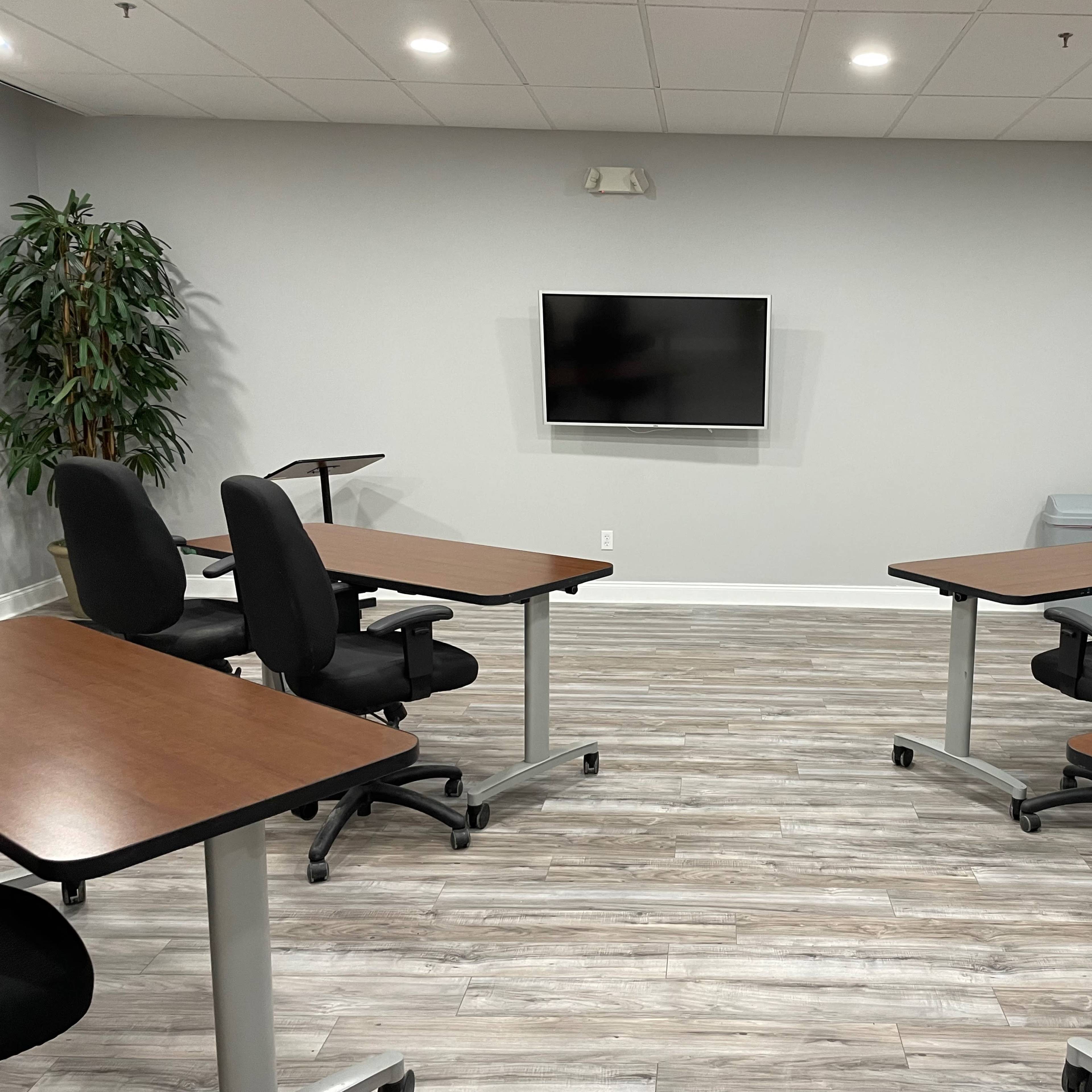 A meeting room features four tables with chairs arranged around them, facing a wall-mounted television, and includes a potted plant in the corner.