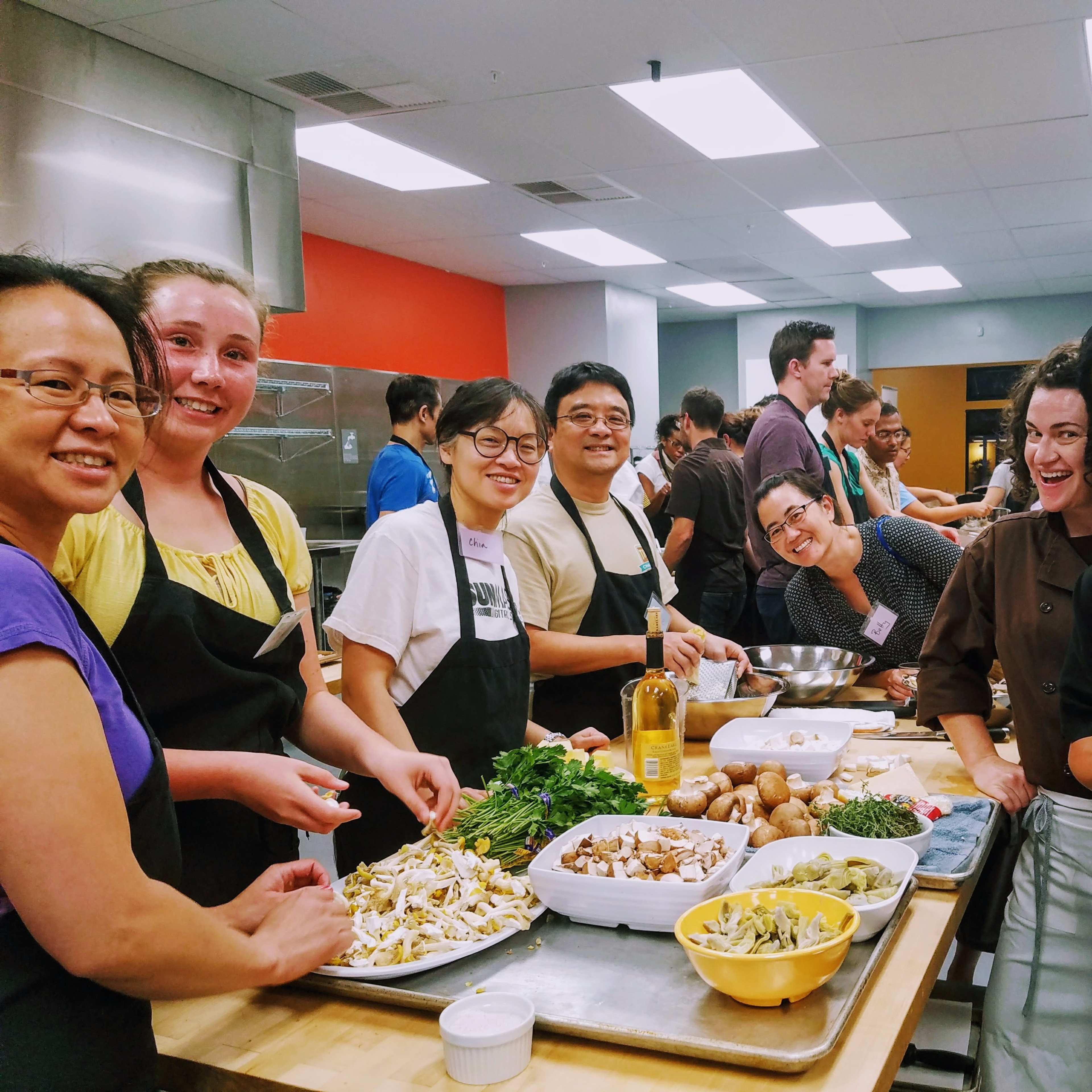 A group of people is preparing food together in a cooking class, standing around a countertop filled with various ingredients and dishes.