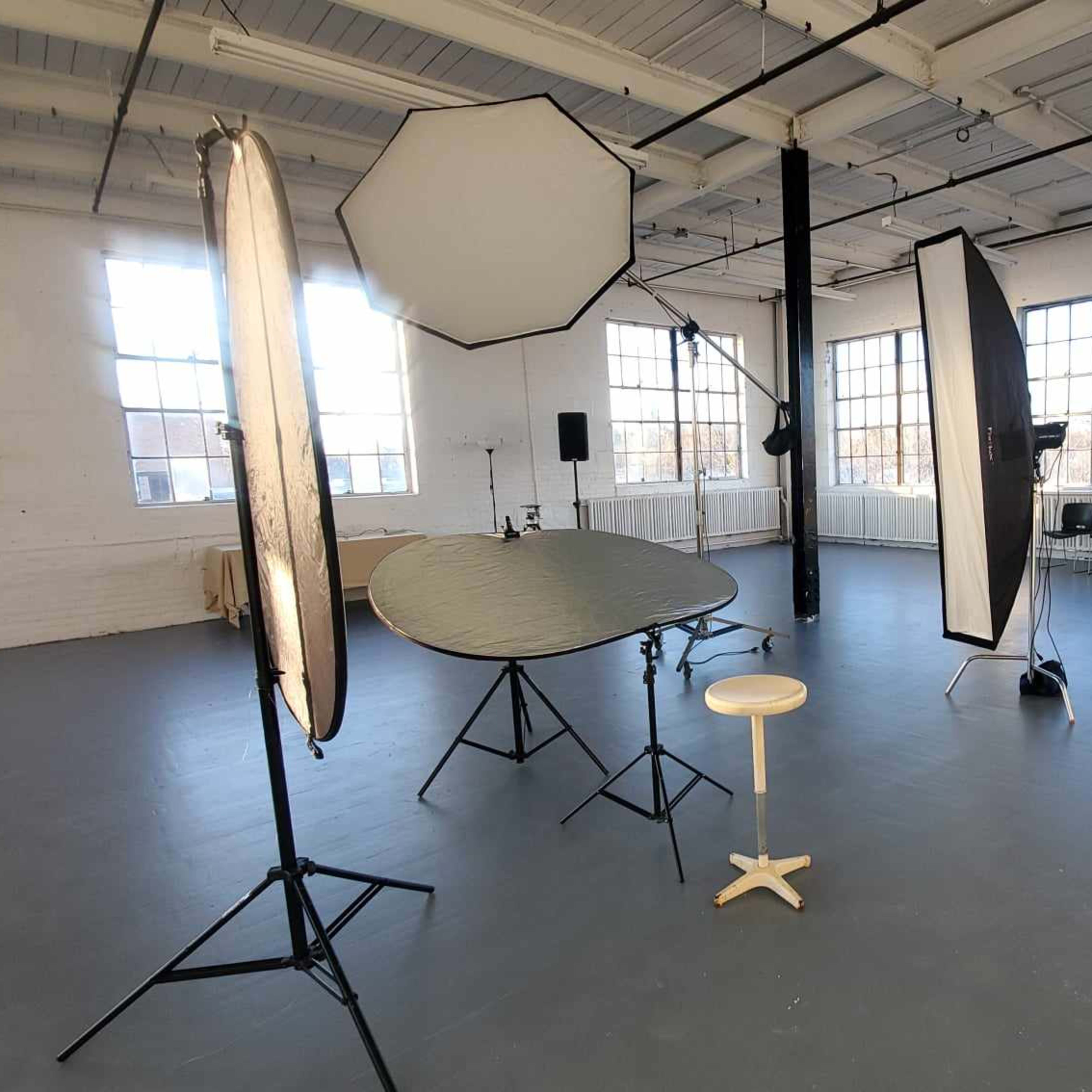 An empty photography studio with various lighting equipment and a stool arranged in a spacious room filled with natural light.