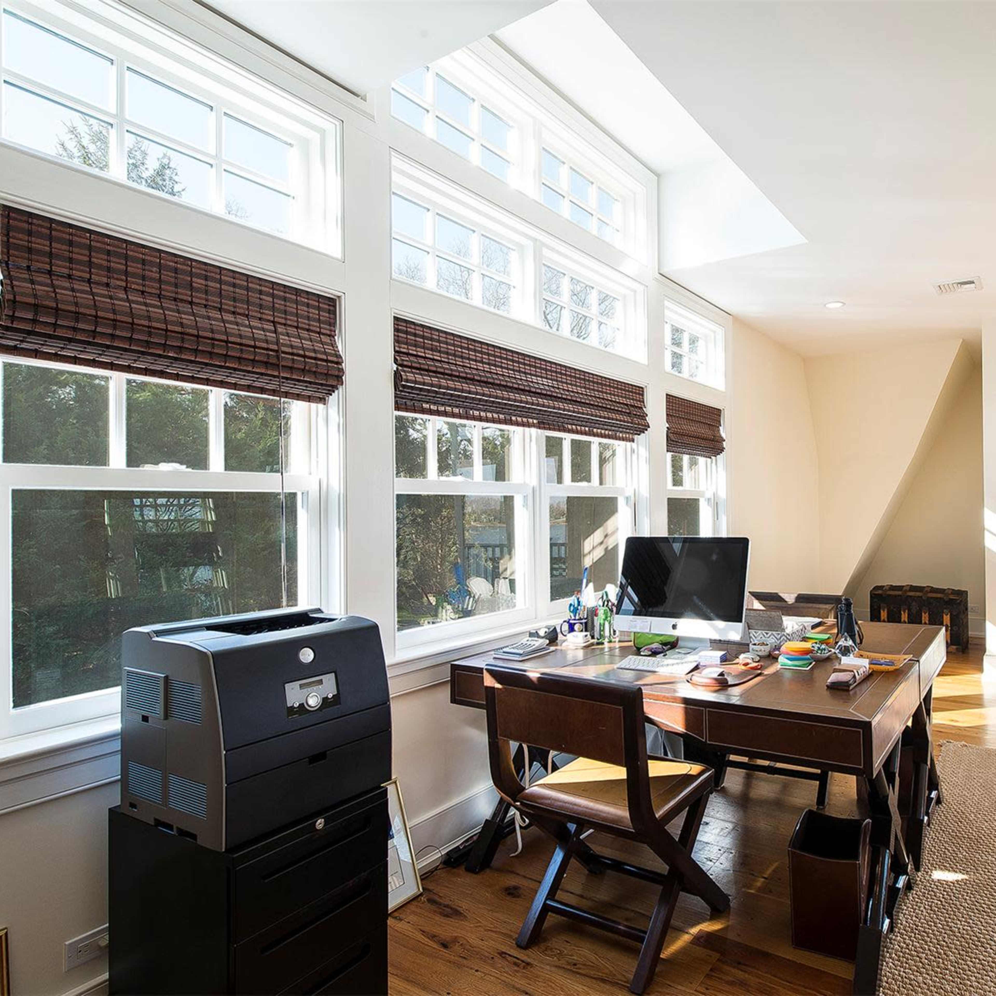 A home office with a wooden desk, a computer, and large windows that allow natural light to enter, facing a view of the outdoors.