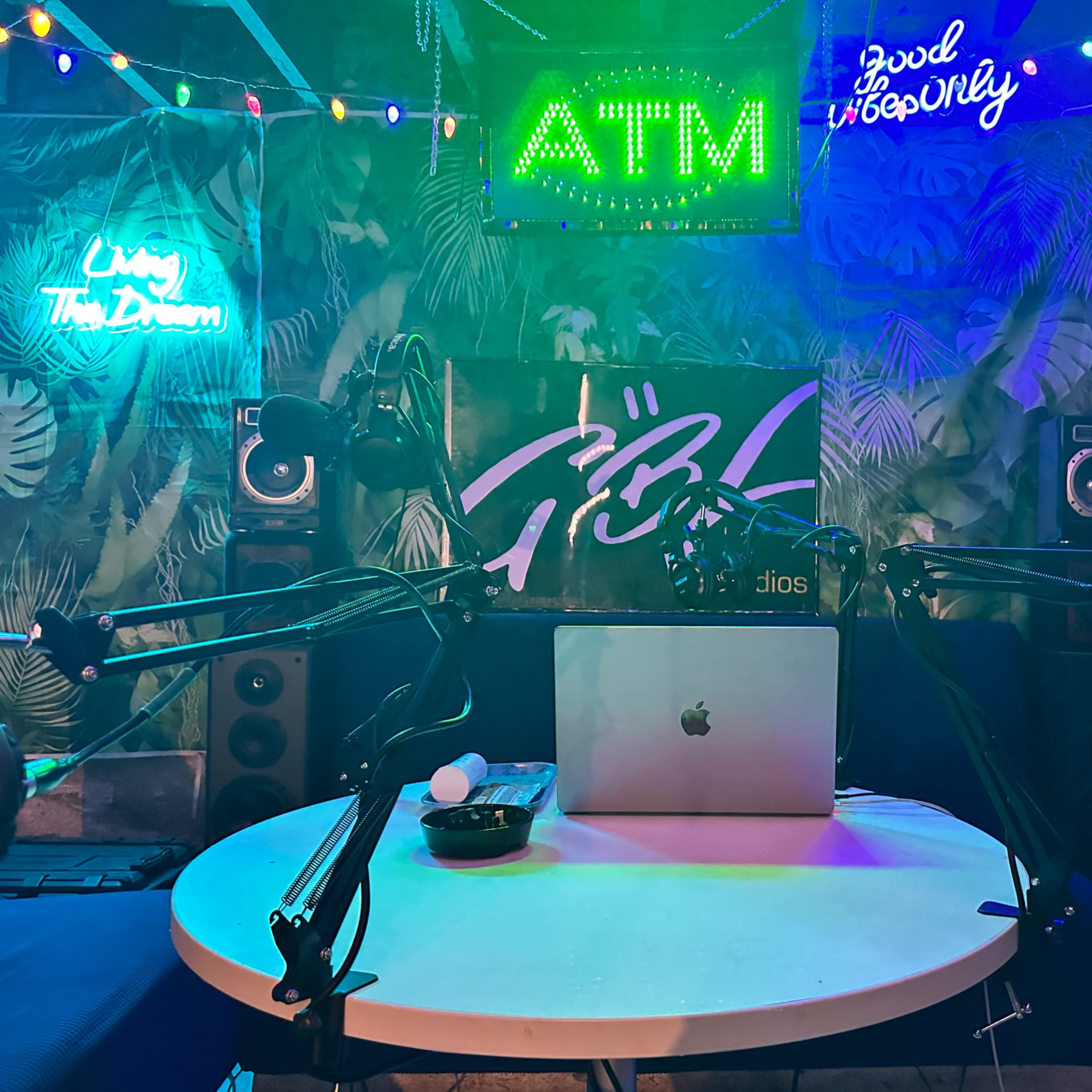 A small studio setup features a table with a laptop, microphone arms, speakers, and colorful neon signs, all surrounded by a vibrant backdrop.