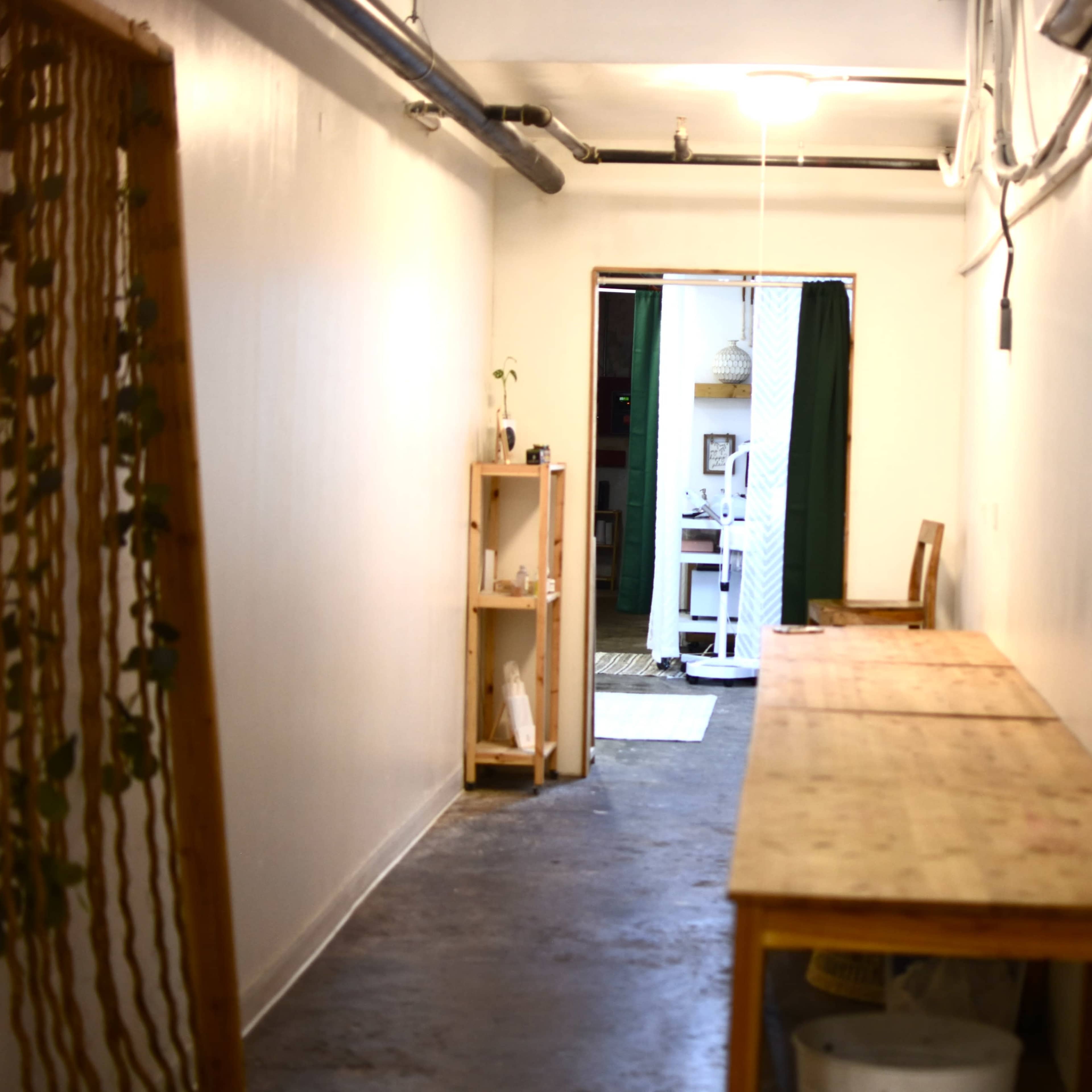 A narrow, well-lit hallway with wooden tables along one side and an open doorway leading to a room at the end.
