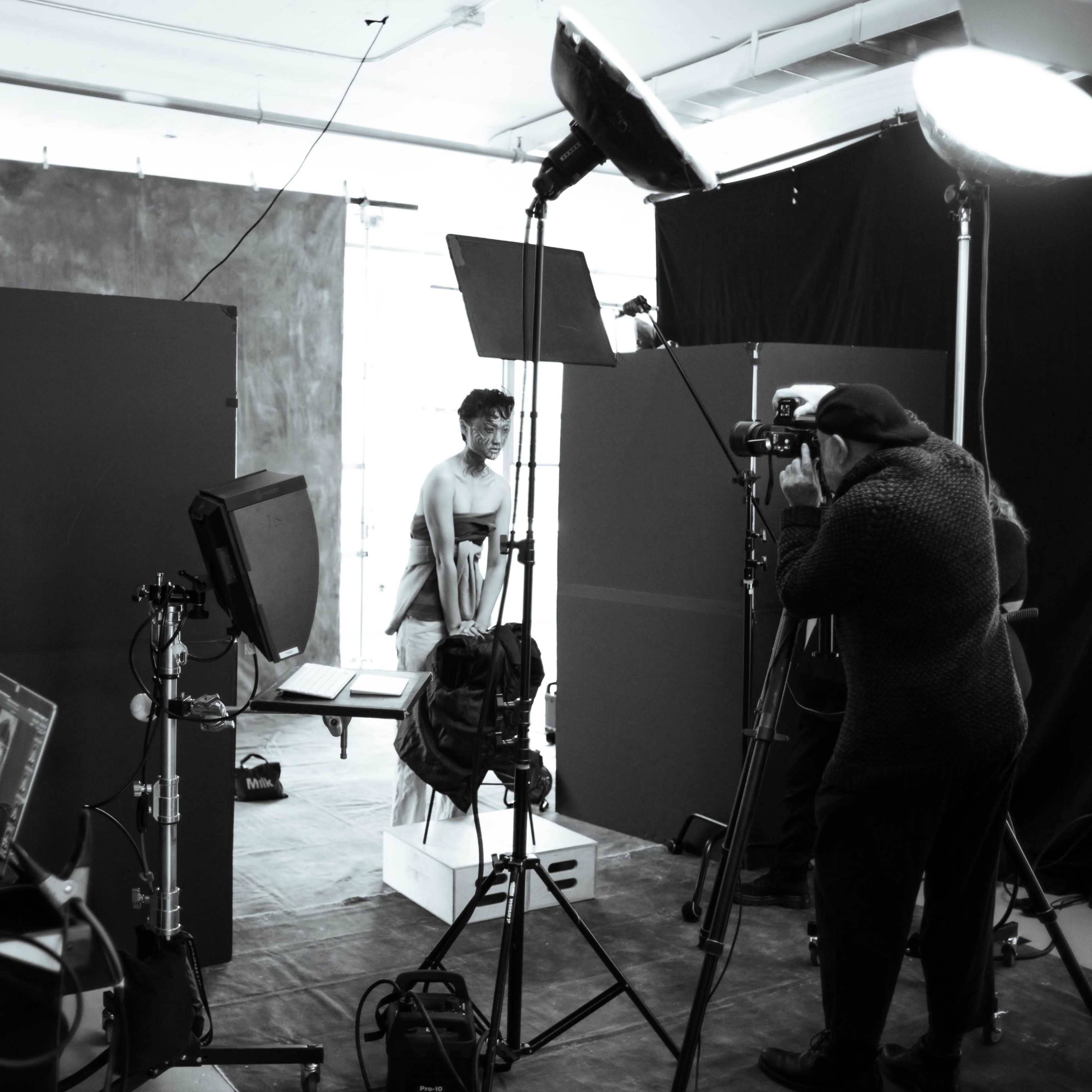 A photoshoot setup features a model posing on a stand, surrounded by lighting equipment and cameras, with a photographer capturing images from a distance.