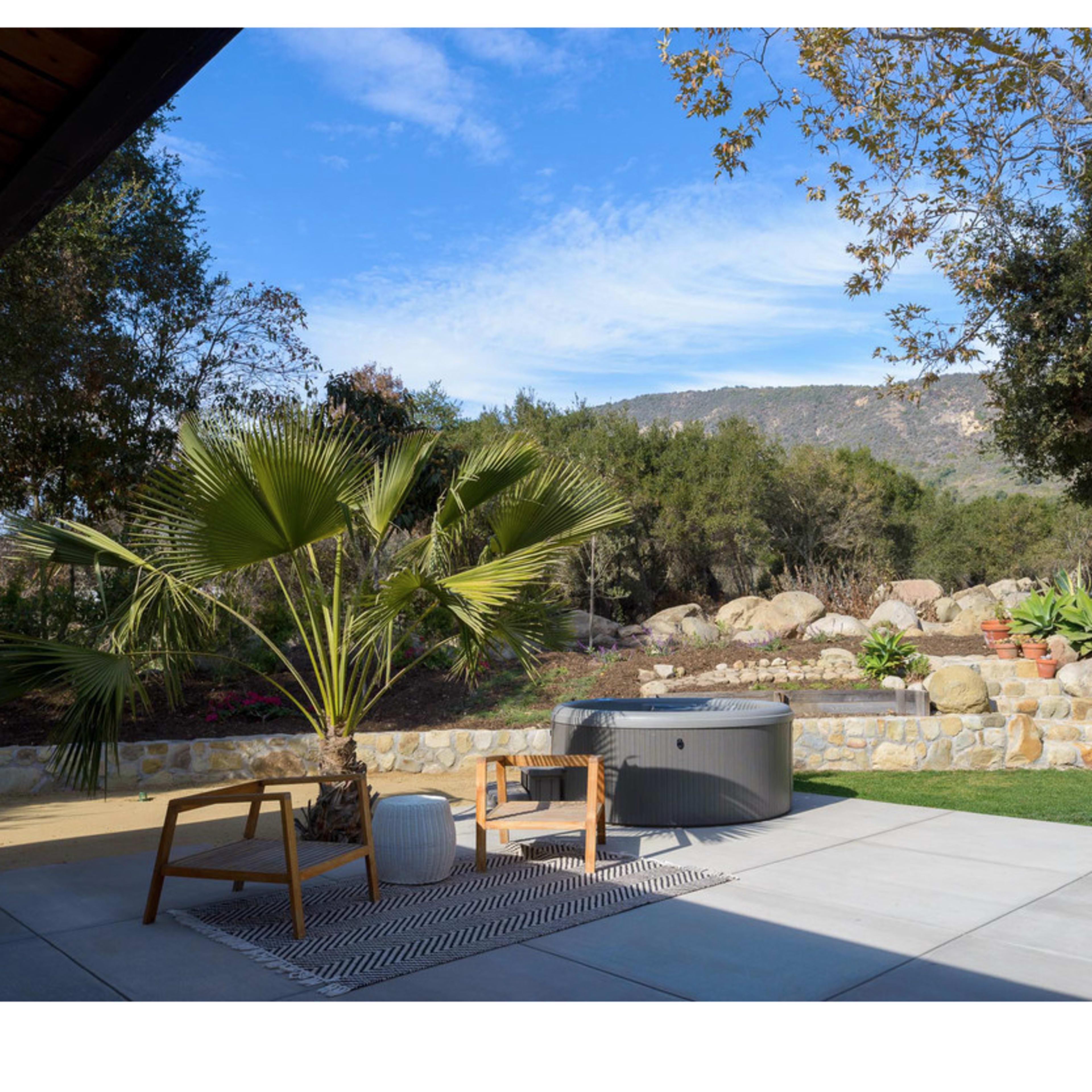 A patio area features a hot tub, a chair, and a palm tree, set against a backdrop of a rocky landscape and distant hills.