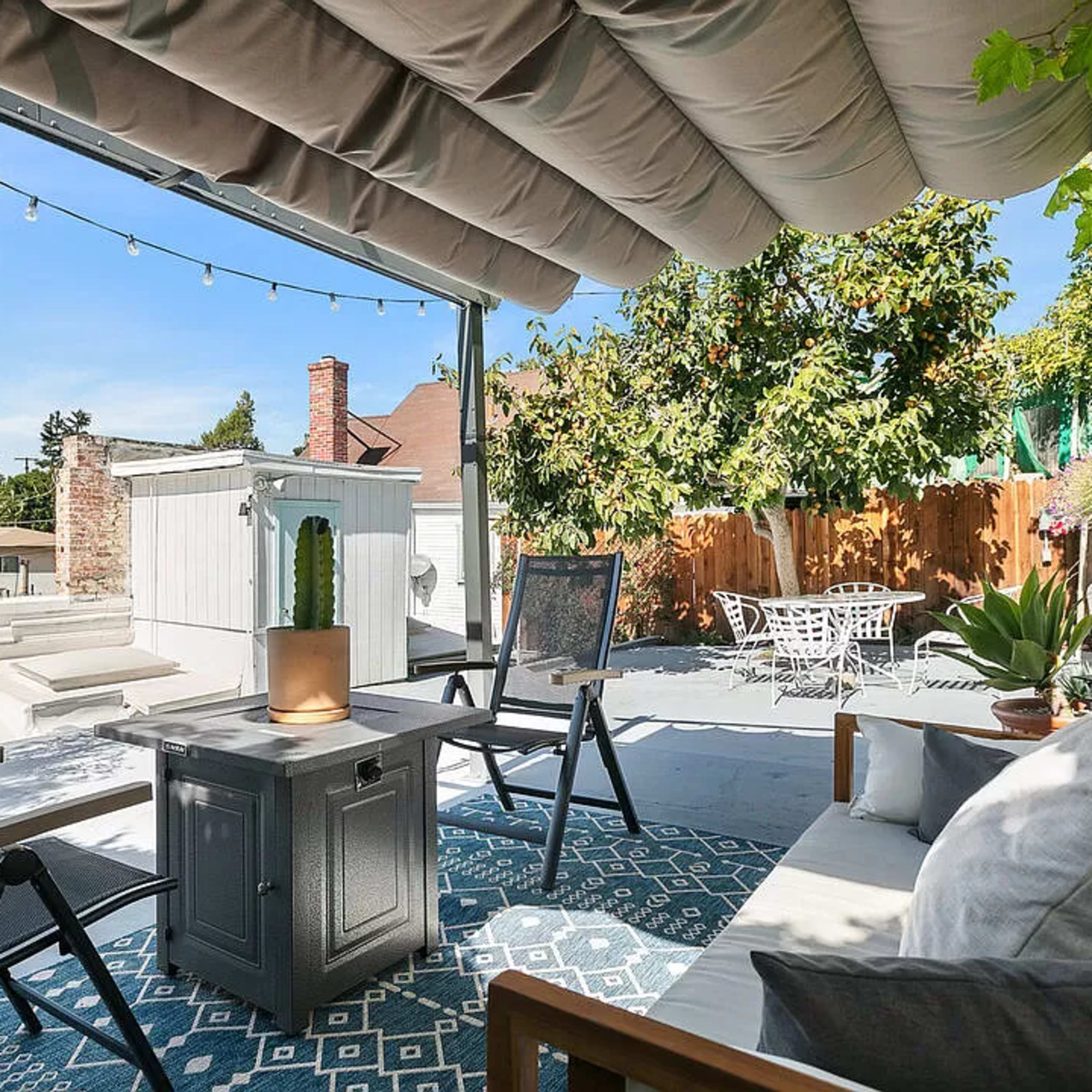 A rooftop patio with outdoor seating, a small table, and potted plants, surrounded by greenery and string lights overhead.