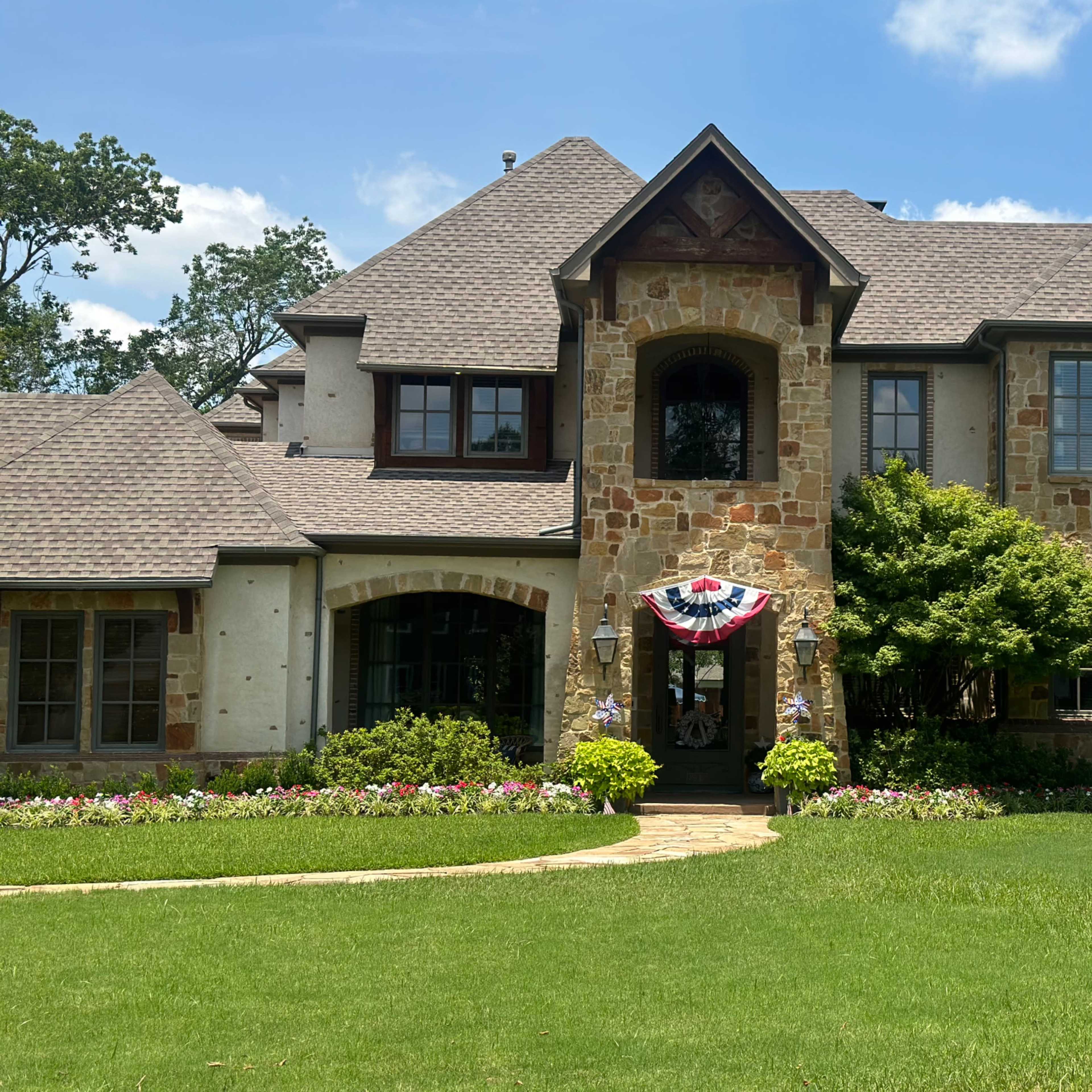 A large, two-story stone and stucco house with a landscaped front yard featuring flower beds and white adirondack chairs.