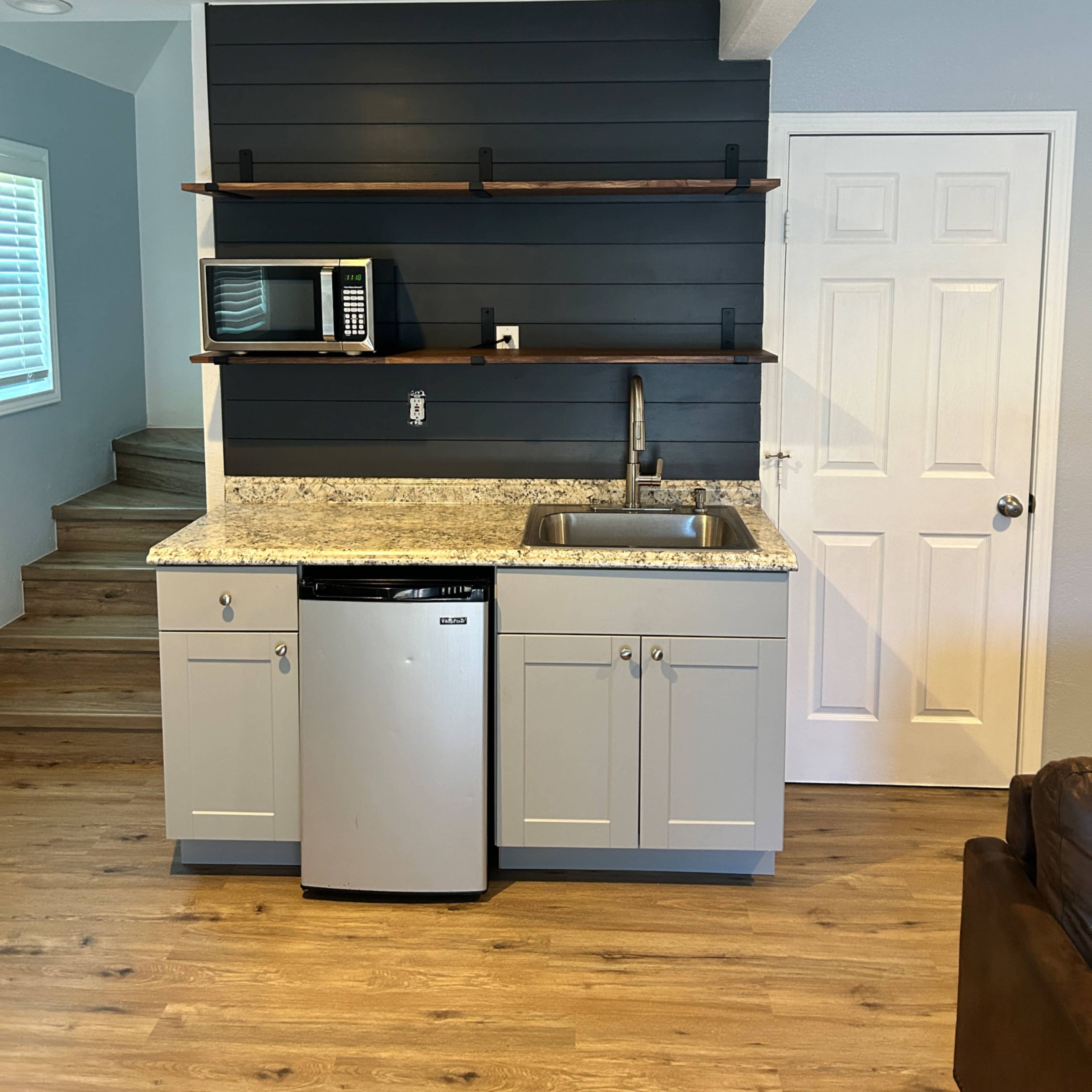 A compact kitchen area with gray cabinets, a stainless steel refrigerator, a sink, a microwave, and wooden shelves against a dark wall.