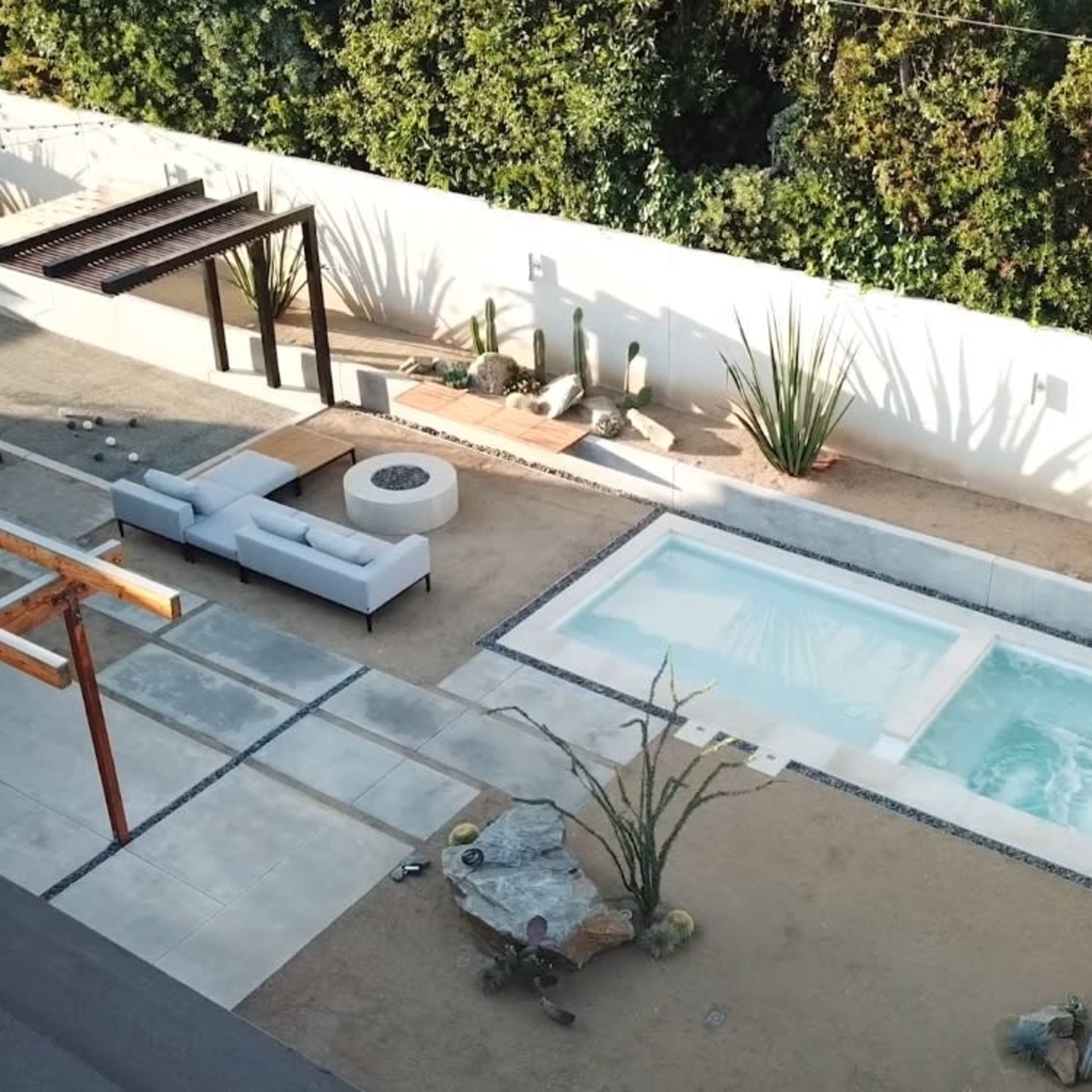 A landscaped backyard featuring a swimming pool, a wooden deck, a lounge area with outdoor furniture, and surrounding plants.