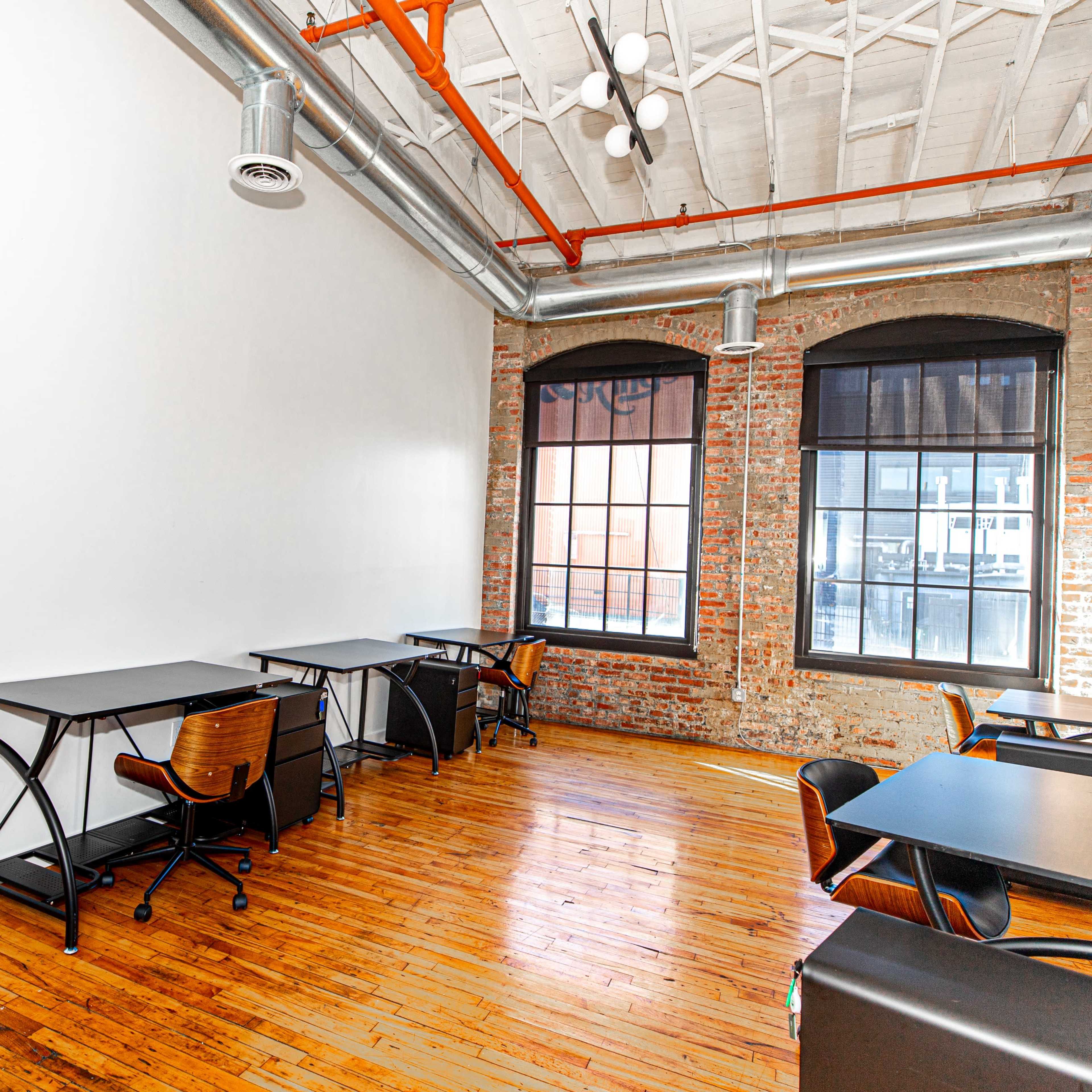 A bright office space featuring two desks with chairs, a black wall board, and large windows that allow natural light to enter.
