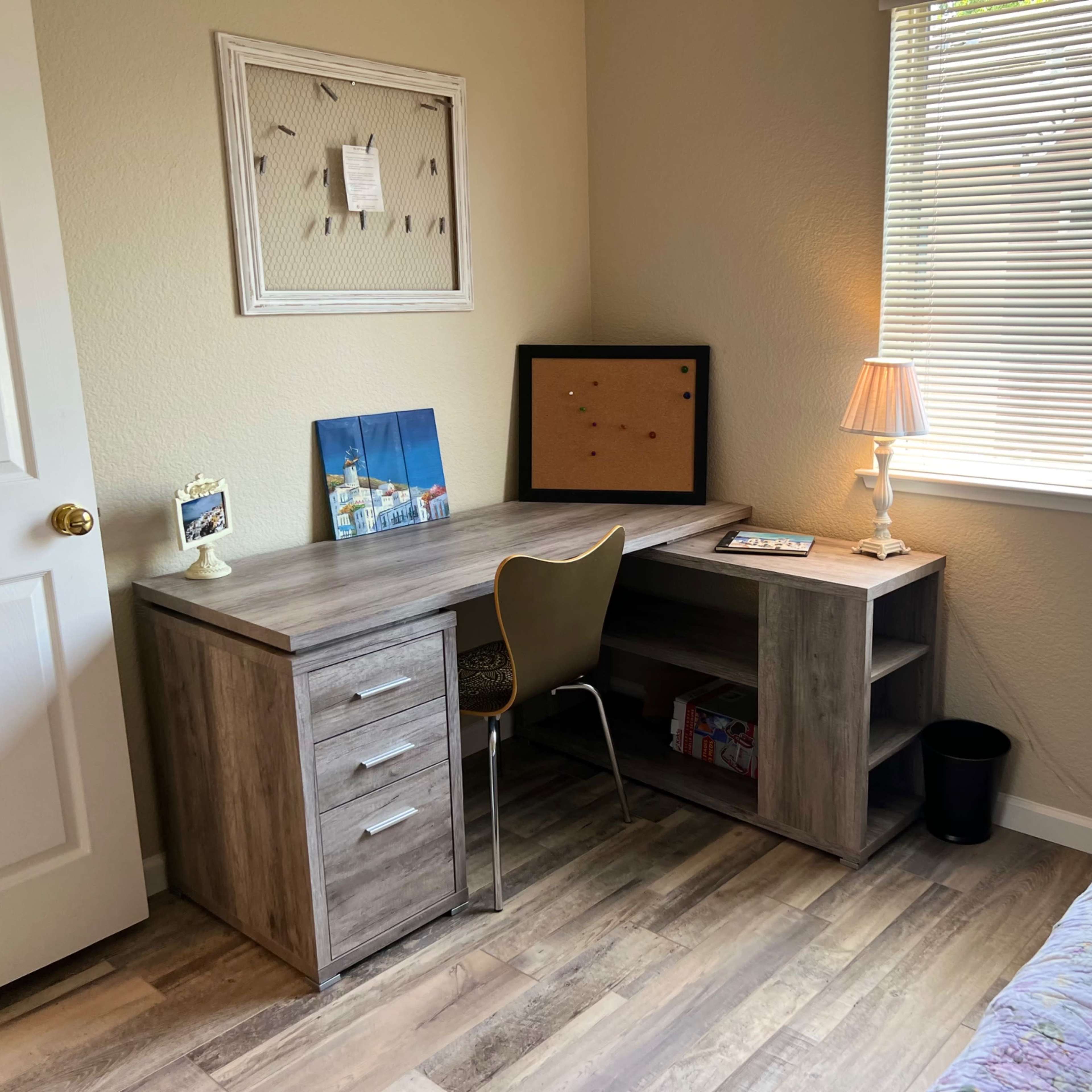 A small, organized workspace featuring a wooden desk, a chair, a lamp, and a bookshelf positioned against a wall with a window.