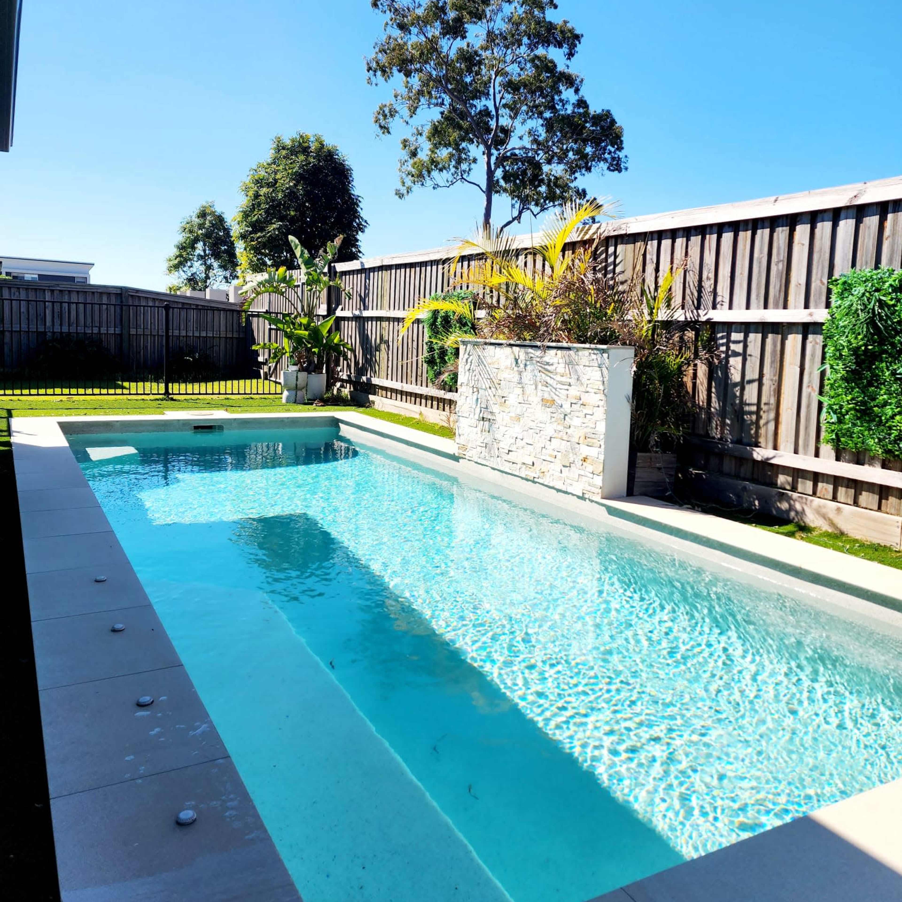 A rectangular swimming pool is surrounded by landscaped greenery and wooden fencing under a clear blue sky.