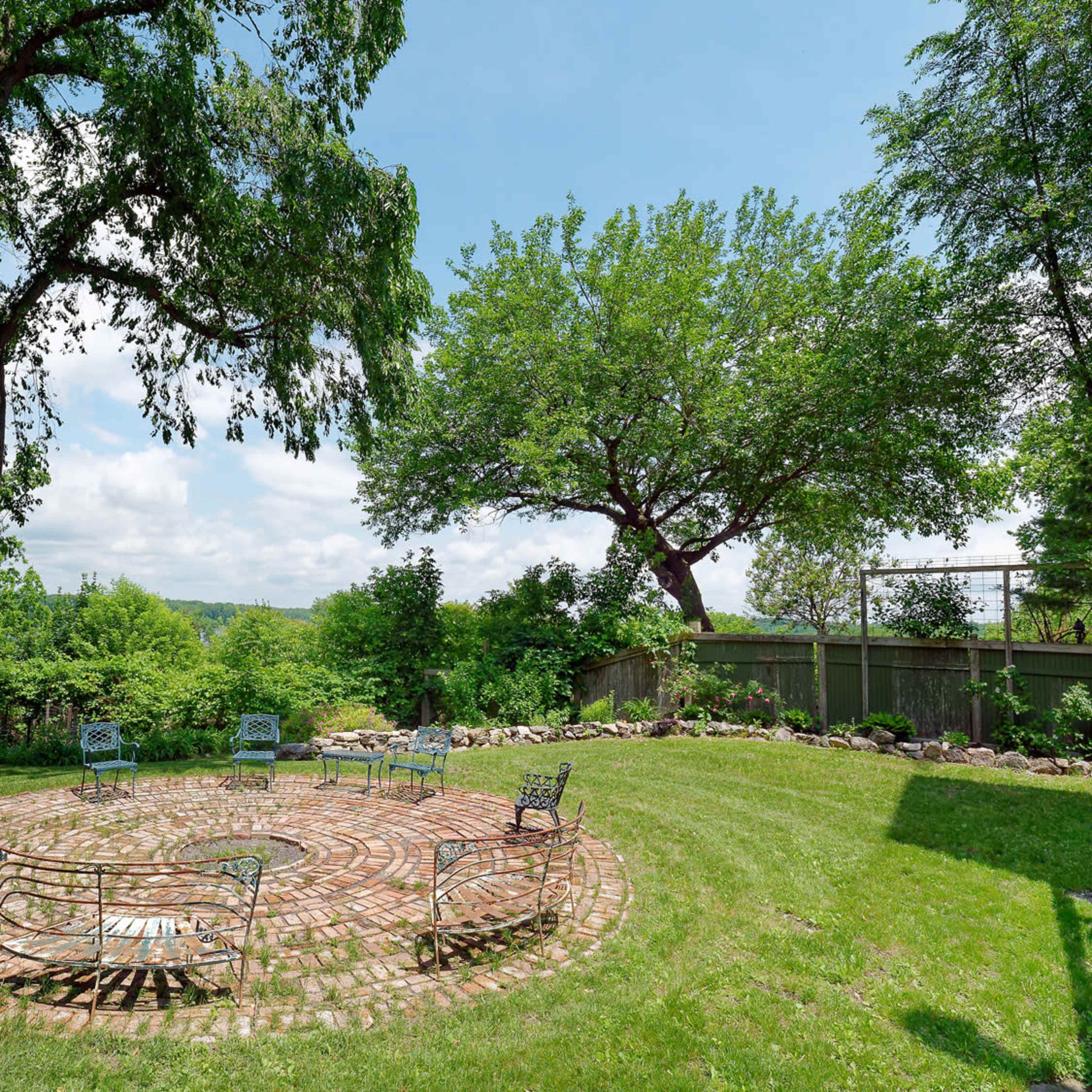 A circular brick seating area with several metal benches is surrounded by grass and trees in a landscaped outdoor space.