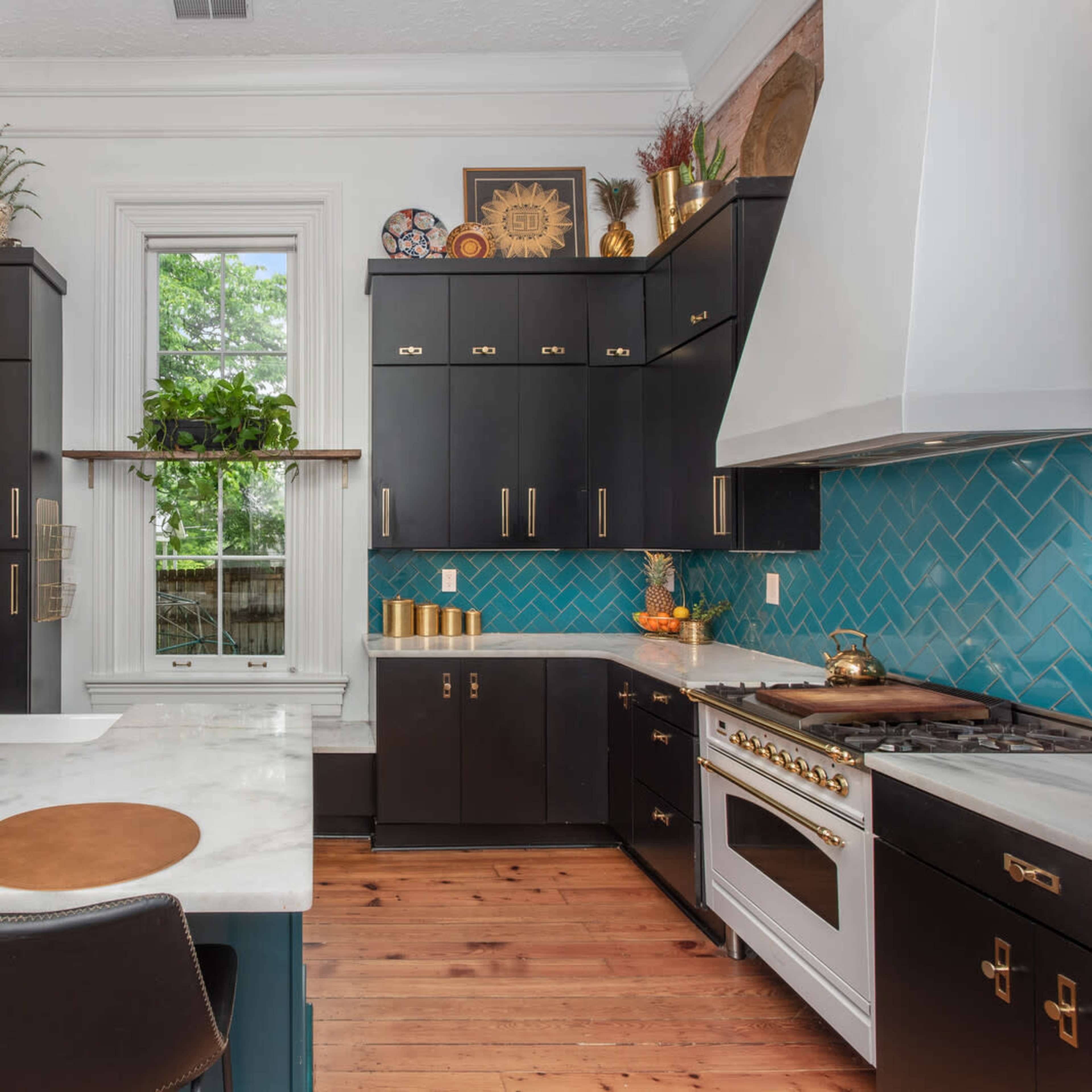 A modern kitchen featuring black cabinetry, a white marble countertop with circular placemats, a teal backsplash, and a large window with greenery outside.