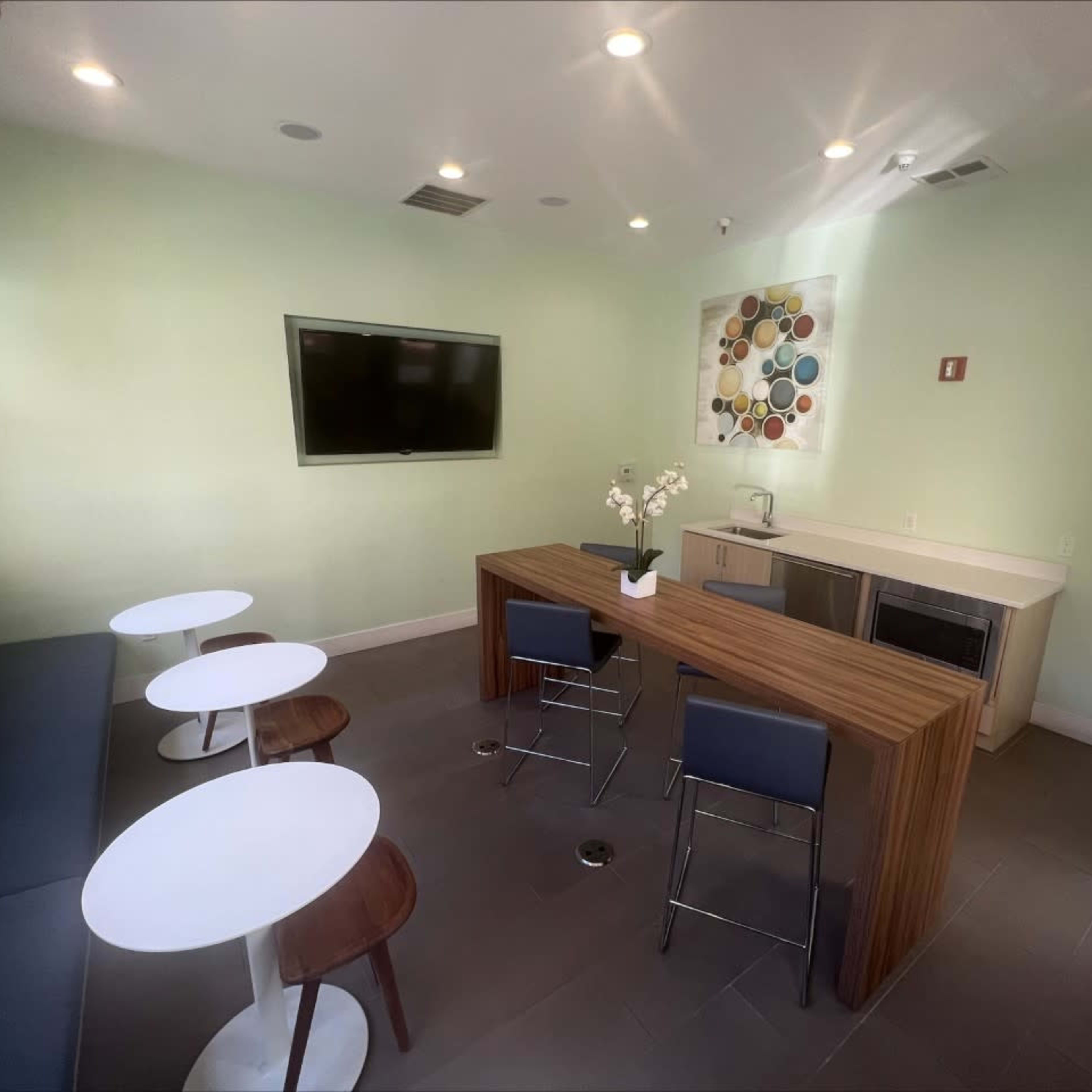 A modern lounge area with a kitchenette, featuring a wooden table and four seating areas consisting of two small round tables and two chairs.