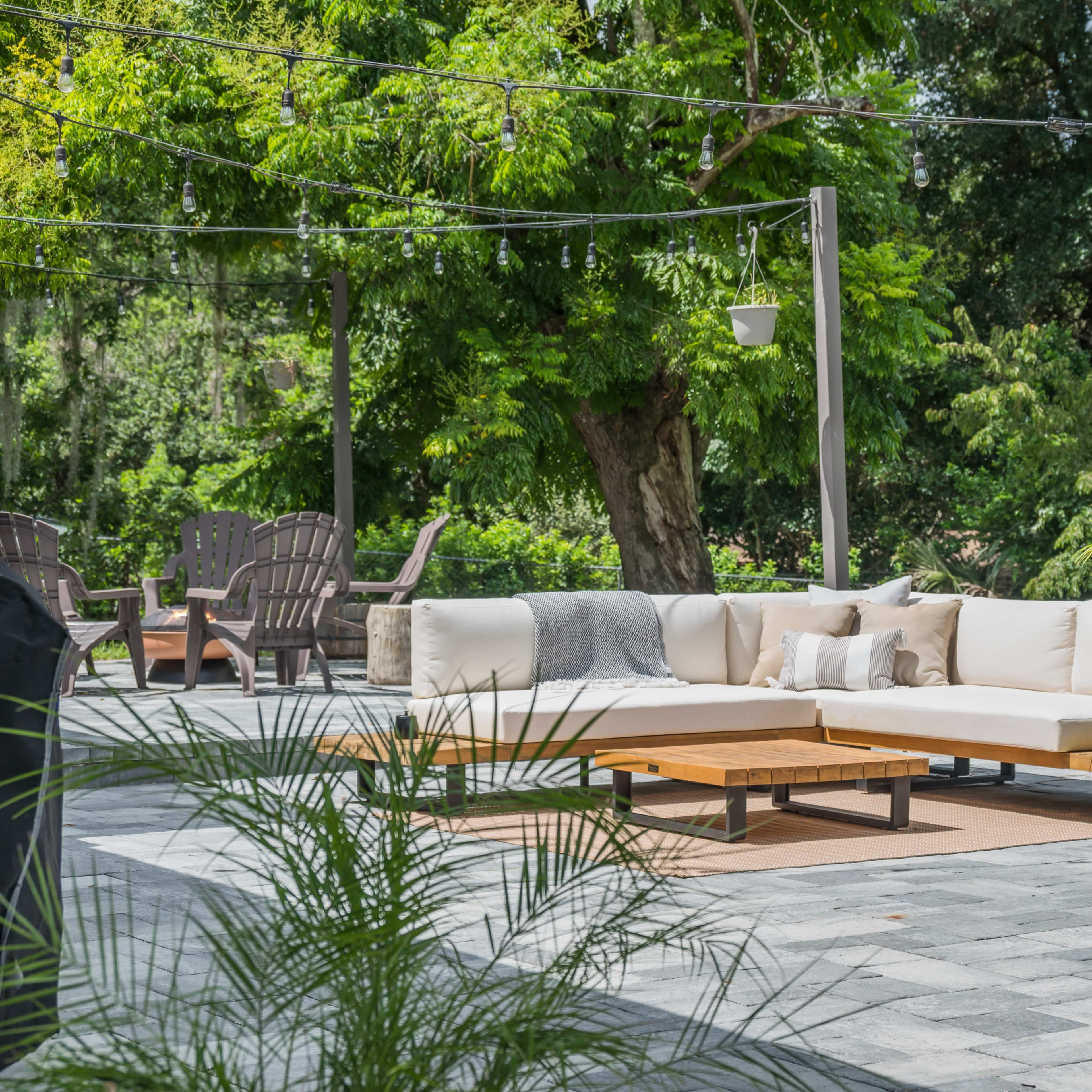 An outdoor seating area with a sectional sofa, wooden tables, and potted plants on a stone patio surrounded by greenery.