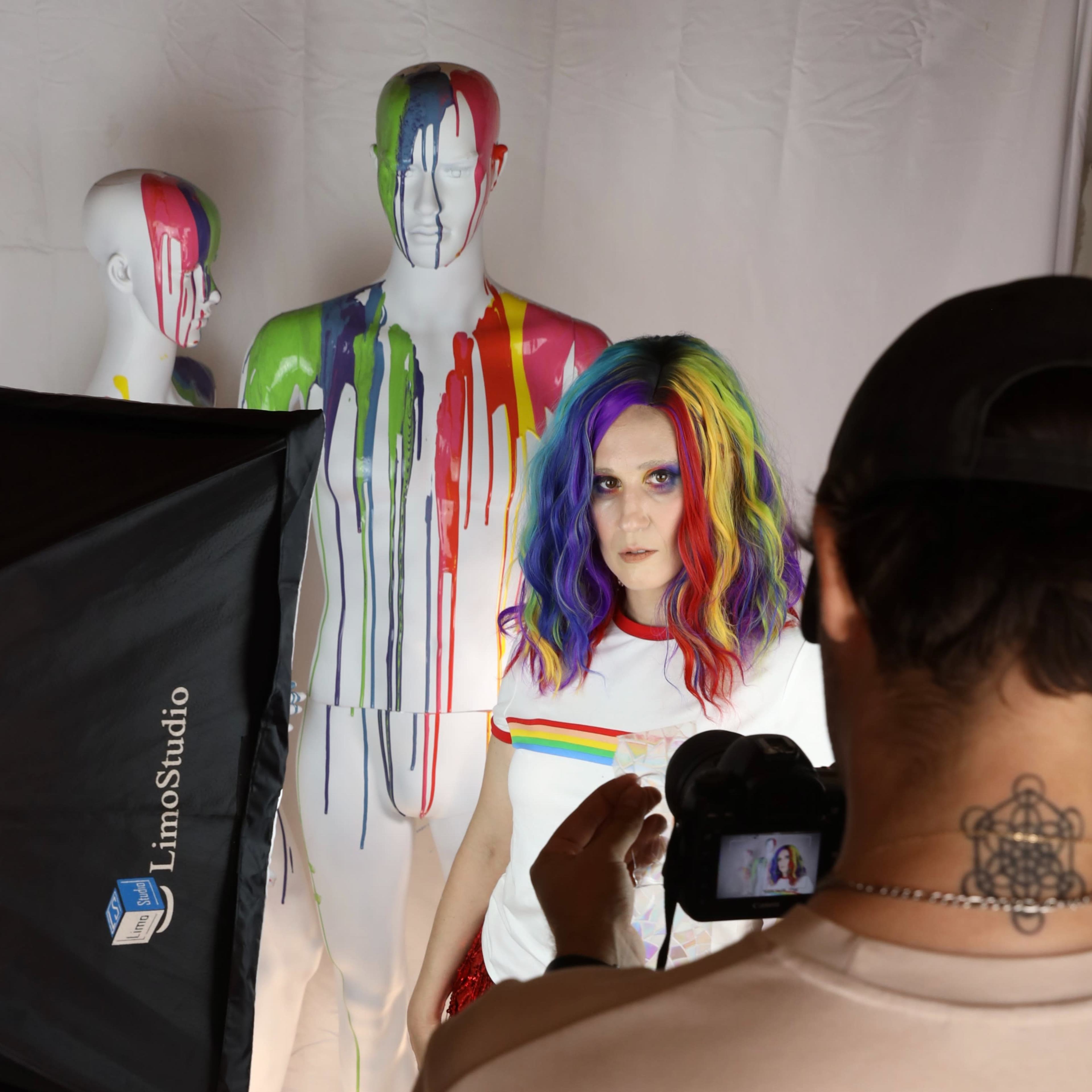 A person with rainbow-colored hair stands in front of two painted mannequins while being photographed against a white backdrop.