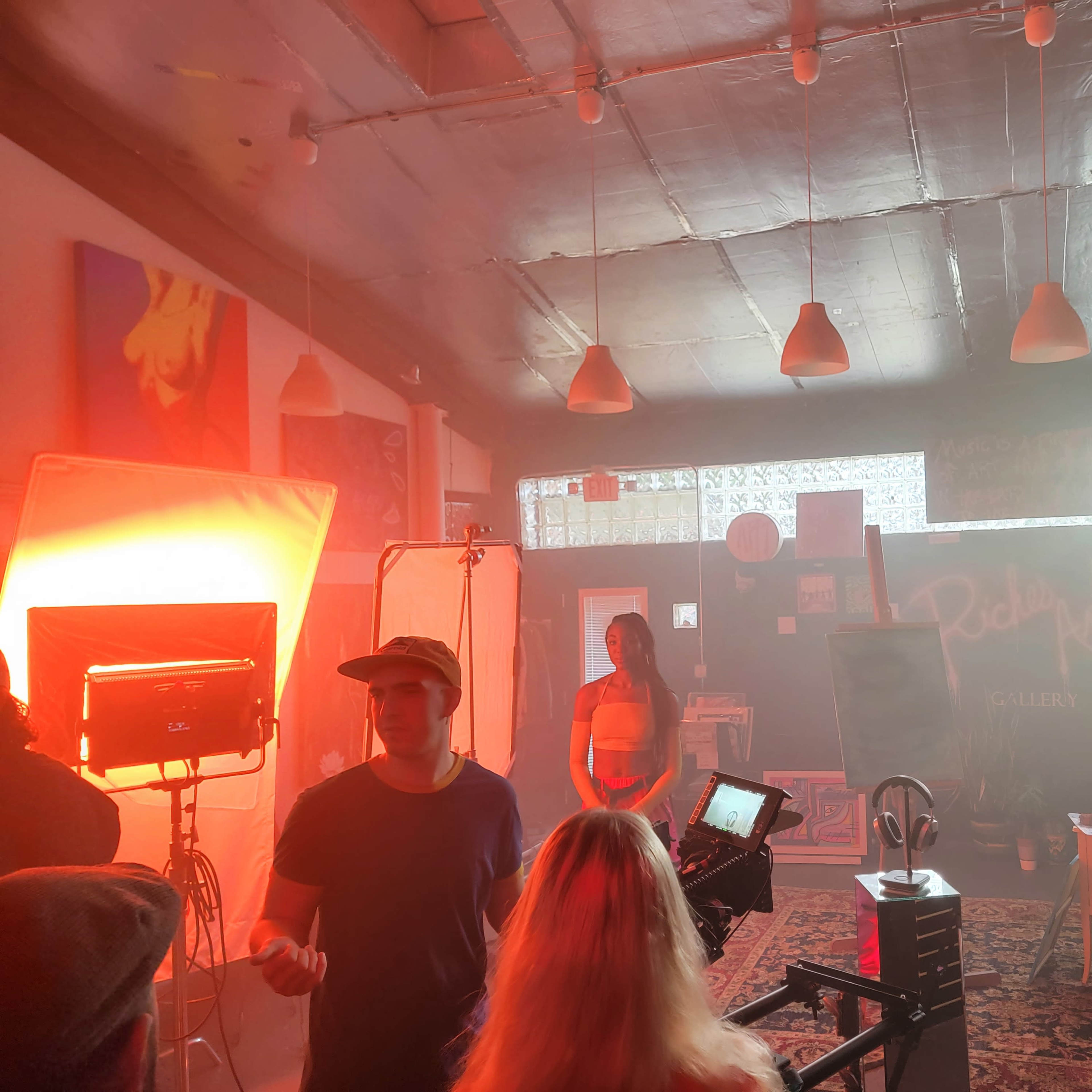 A well-lit gallery scene features a model posing in front of a backdrop, surrounded by production equipment and crew members.