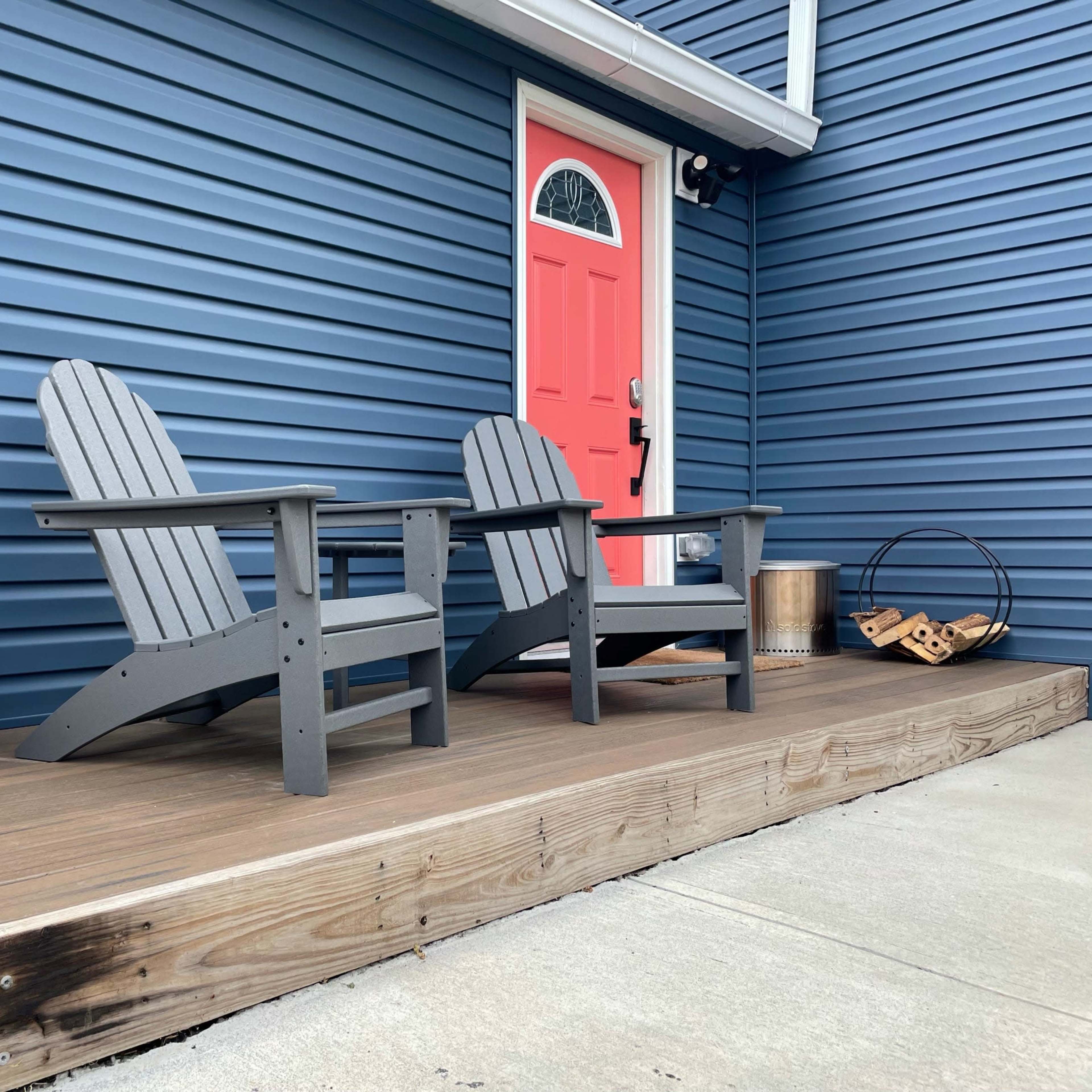 Two gray Adirondack chairs positioned on a wooden deck in front of a blue house with a pink door and a red planter.