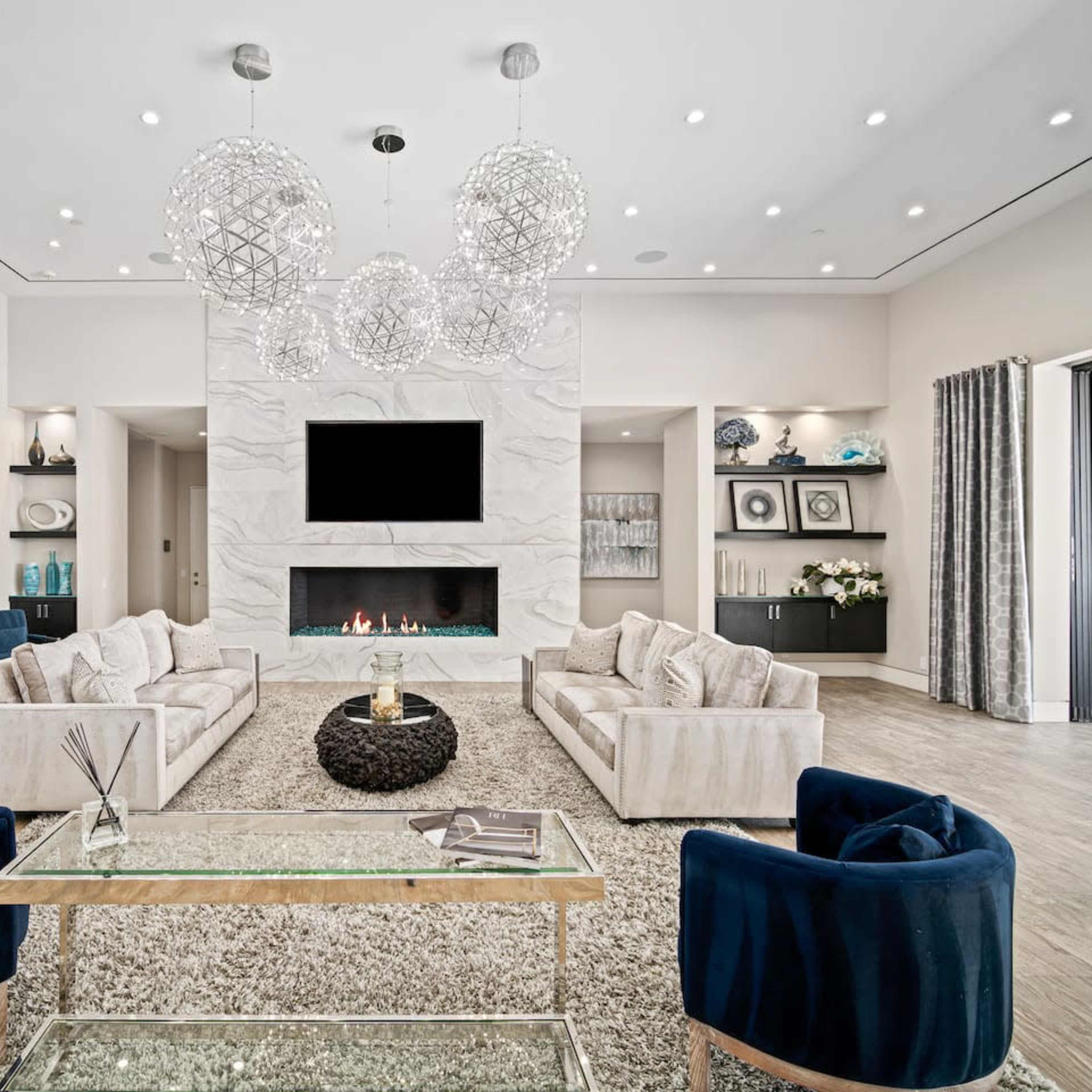 A spacious modern living room features two large sofas facing each other, a central coffee table, and decorative lighting, with a television mounted above a sleek fireplace.