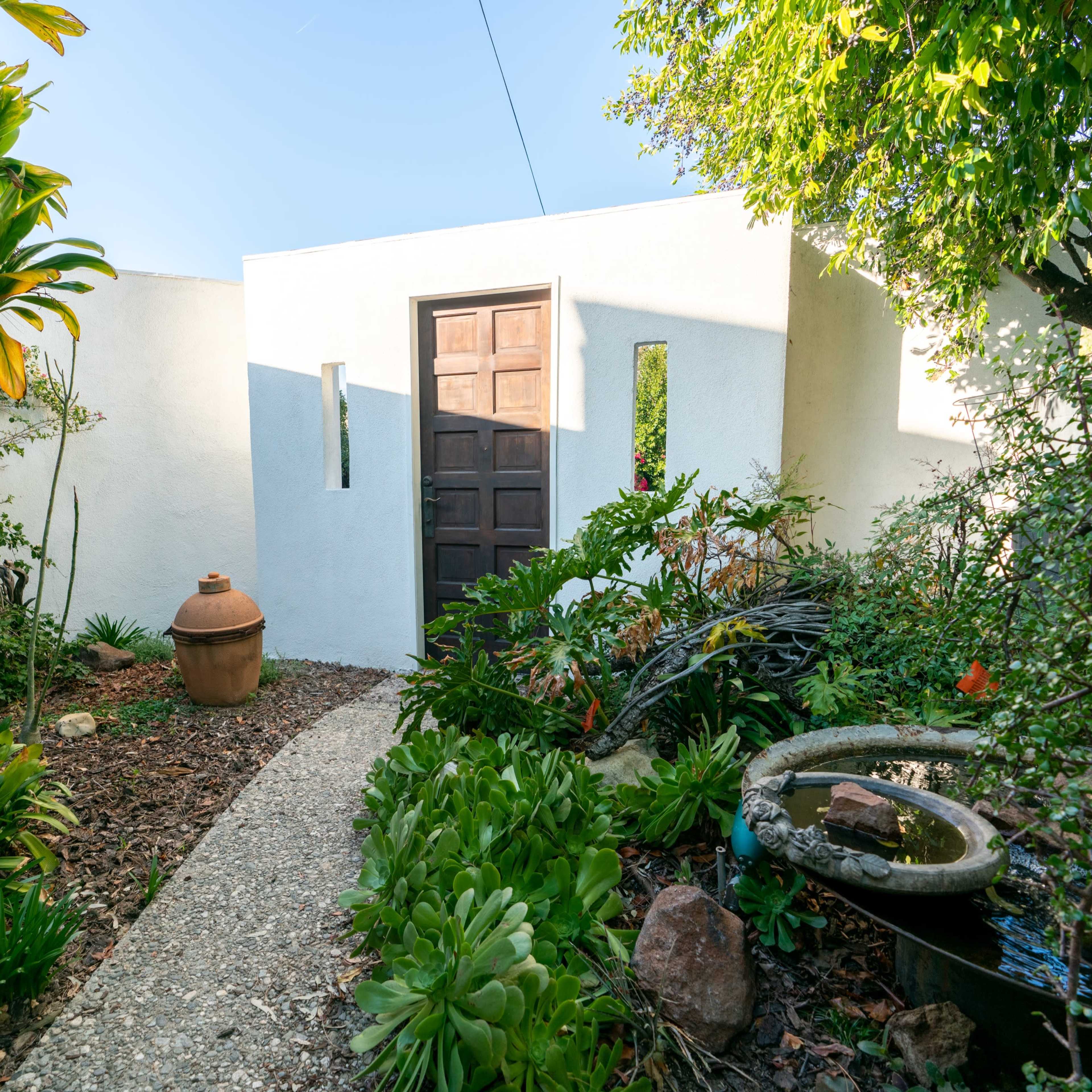 A pathway lined with greenery leads to a wooden door set into a white stucco wall, surrounded by various plants and a decorative fountain.