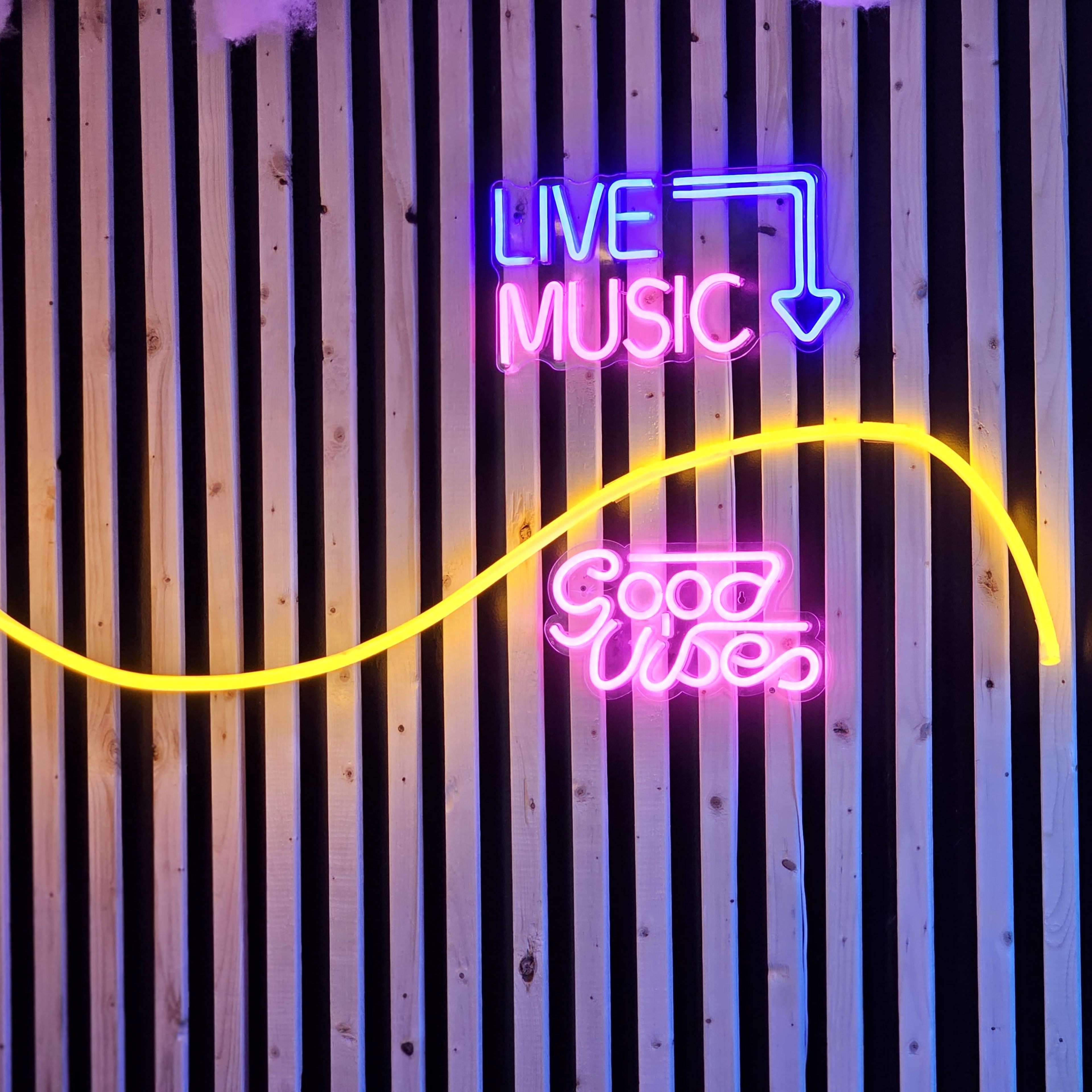 A wooden wall adorned with neon signs that read "LIVE MUSIC" with an arrow pointing down and "Good Vibes" in a wavy design.