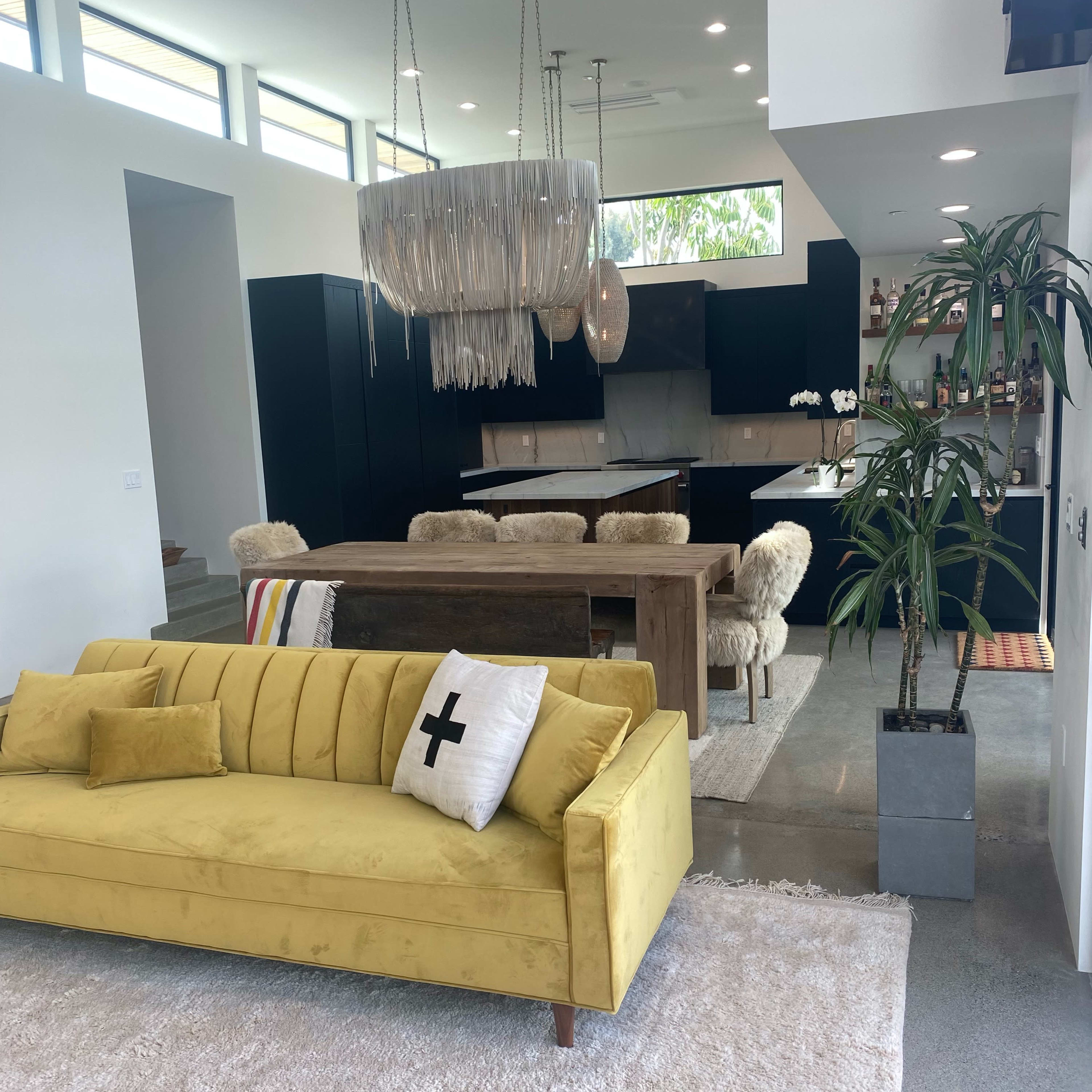 A modern living space featuring a yellow sofa in the foreground, a wooden dining table with fur-covered chairs in the background, and large windows near the ceiling.