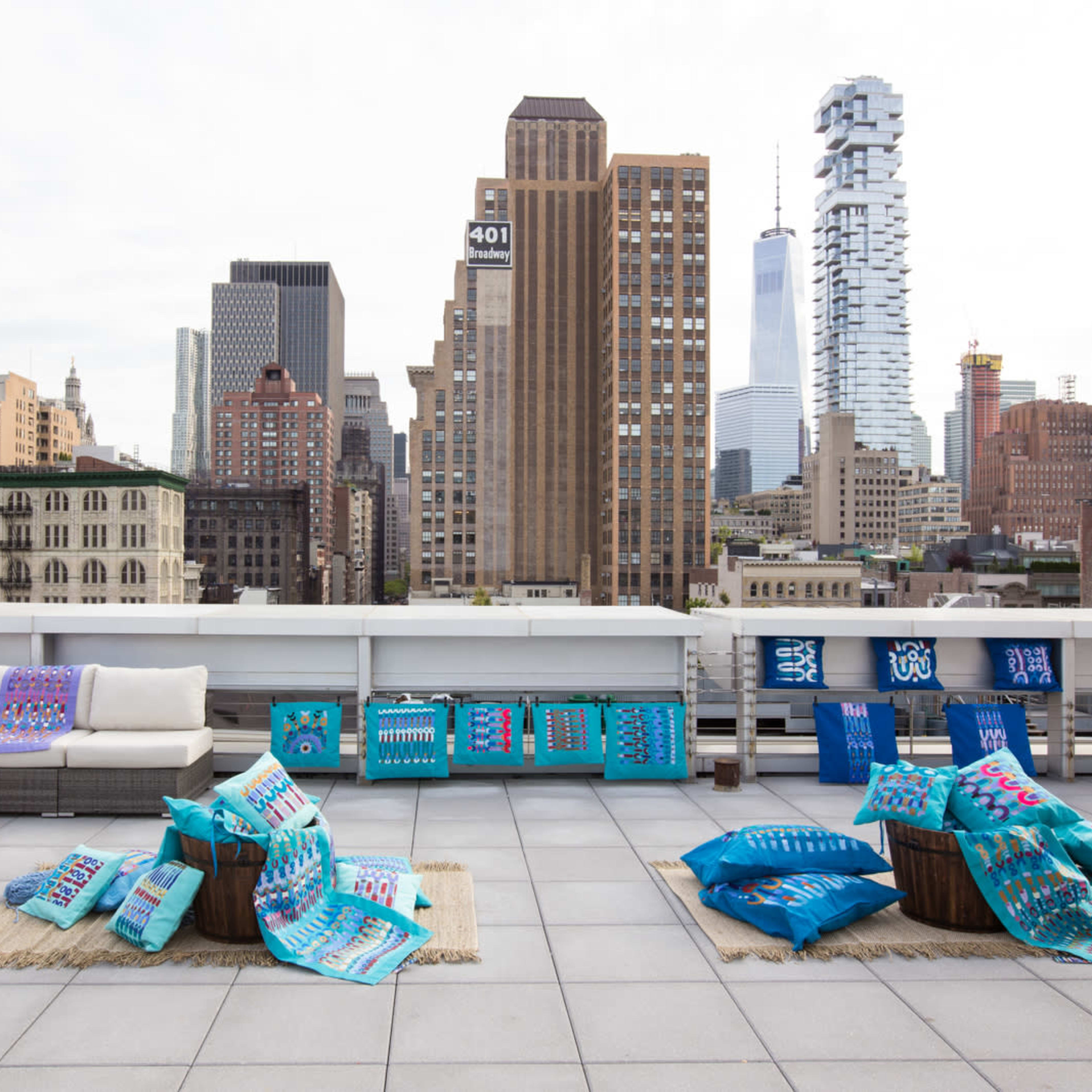 A rooftop space features colorful cushions and throws arranged on the ground, with a view of tall buildings and the skyline in the background.