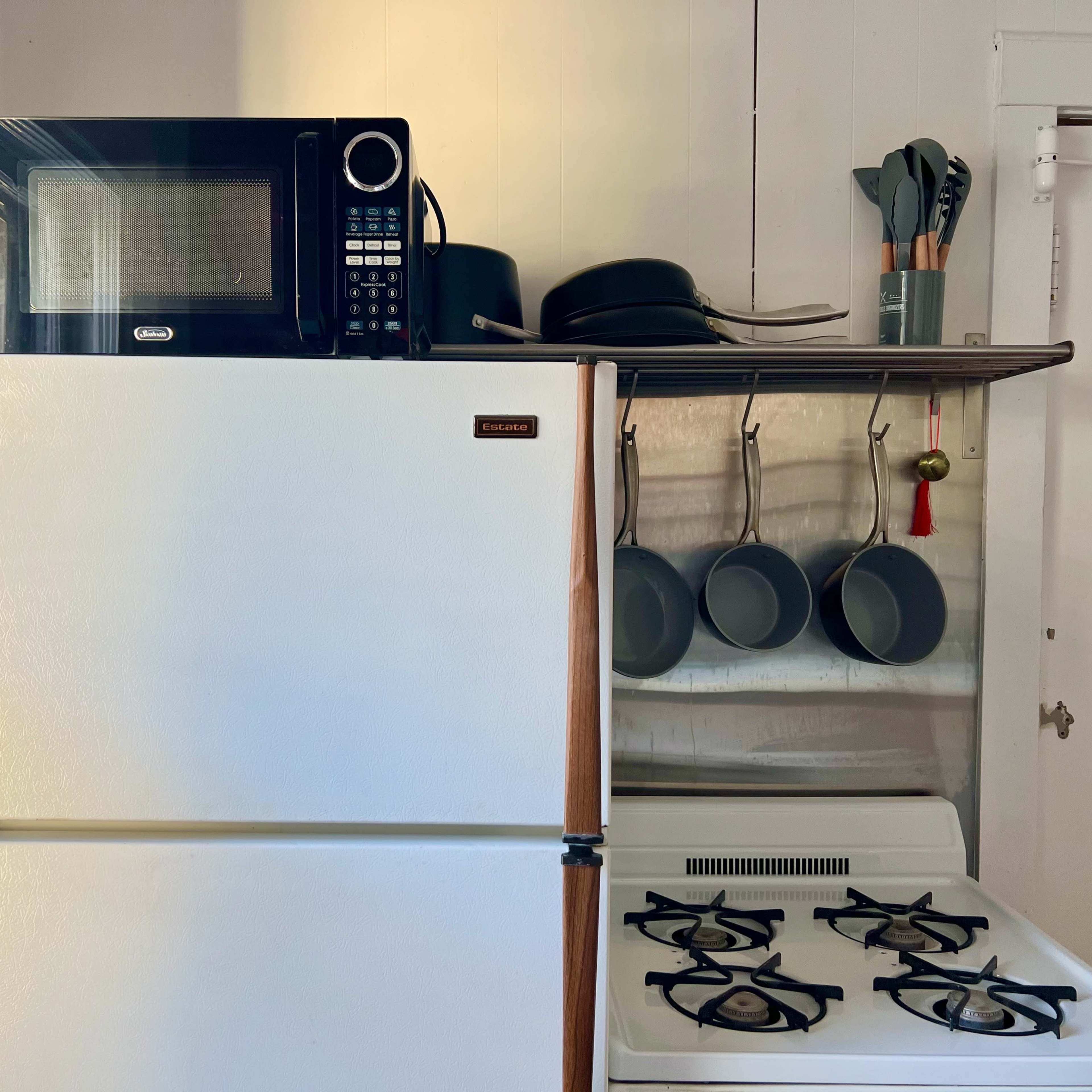 A kitchen setup featuring a white refrigerator topped with a microwave, next to a stove with four burners and a shelf holding several pots and cooking utensils.