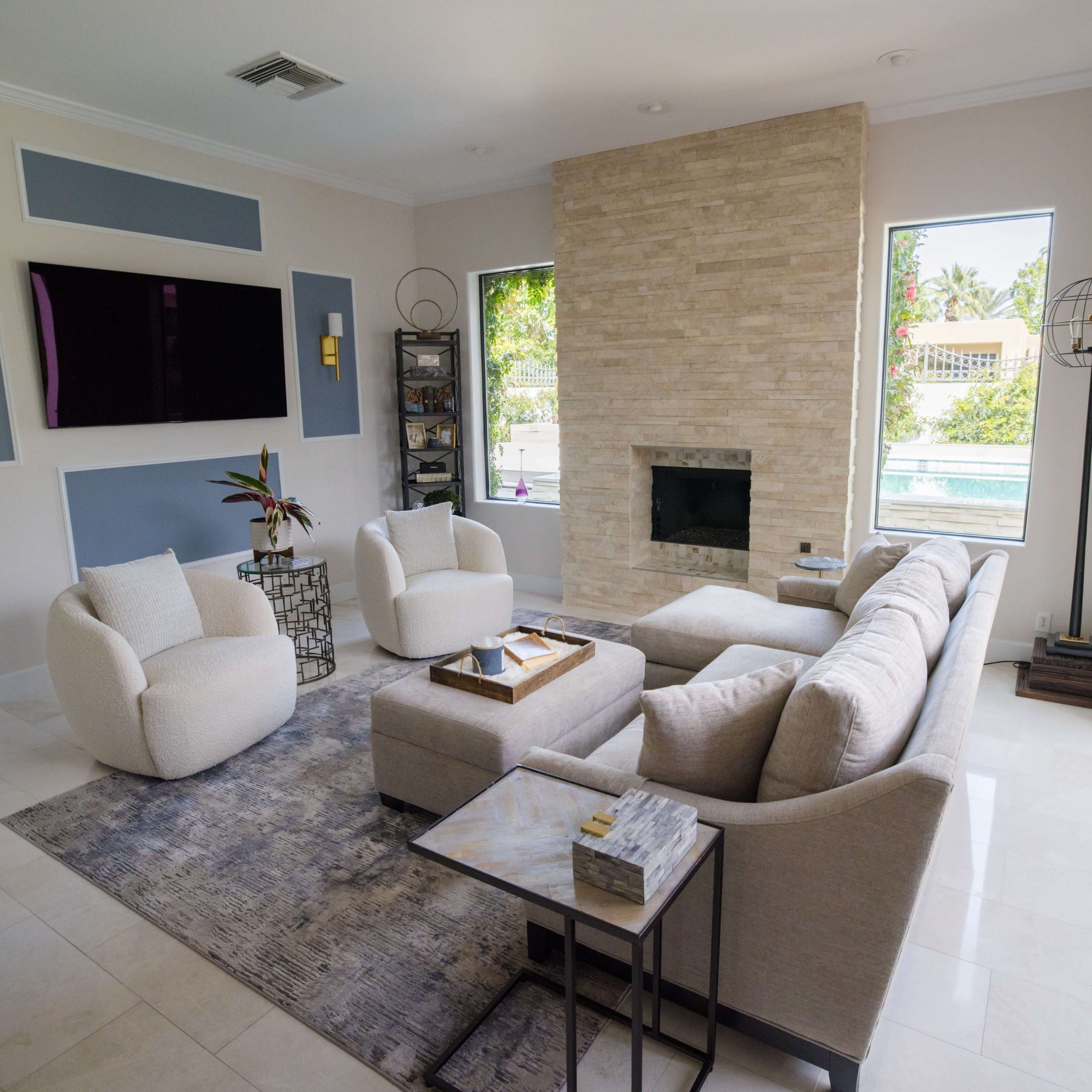 A modern living room featuring a sectional sofa, a coffee table, a stone fireplace, and large windows overlooking a pool area.