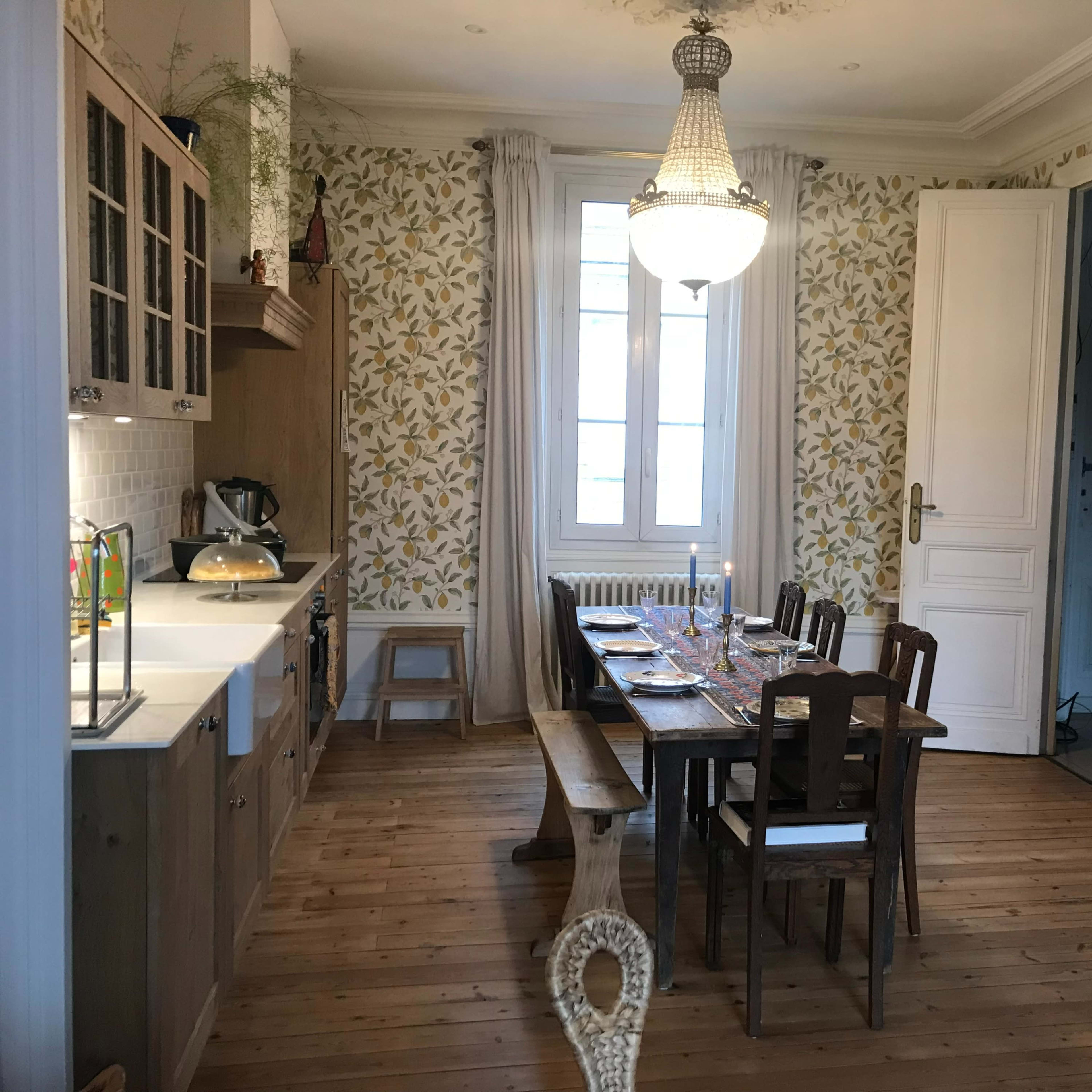 A light-filled kitchen features wooden cabinets, a dining table set for a meal, and floral wallpaper.