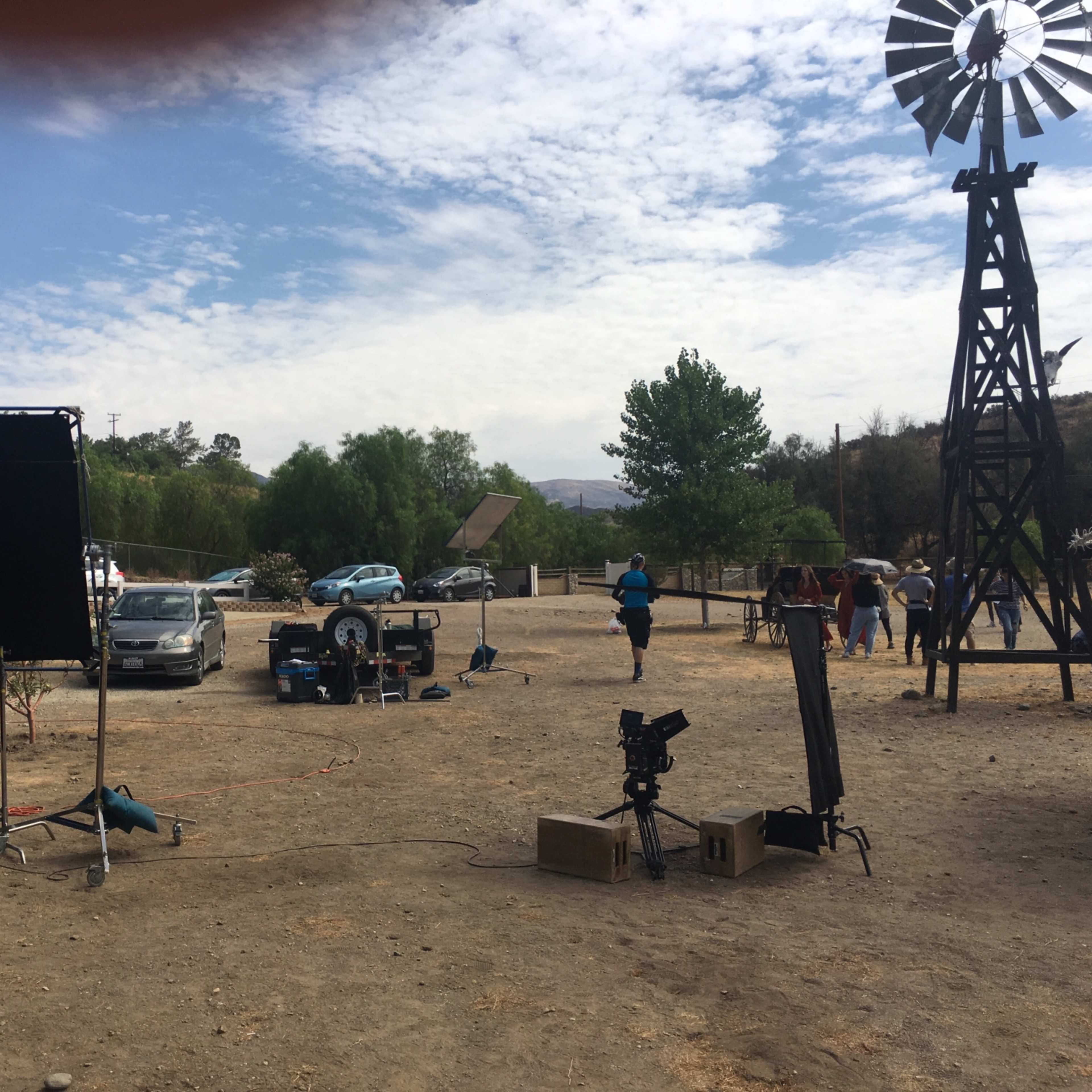 A film crew sets up equipment in a dusty outdoor location with a windmill in the background and vehicles parked nearby.