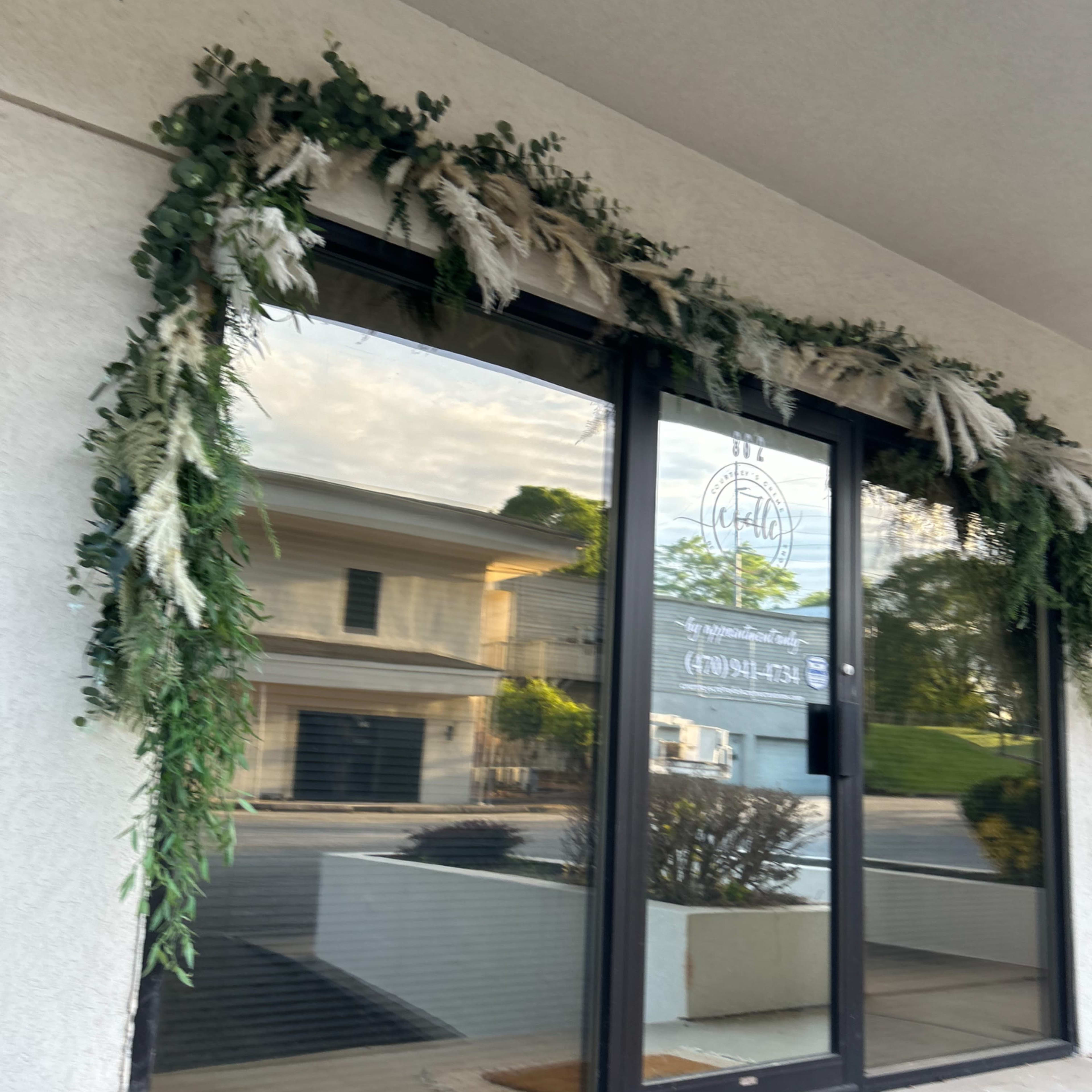 The exterior of a storefront features large glass doors adorned with a greenery and dried flower garland.