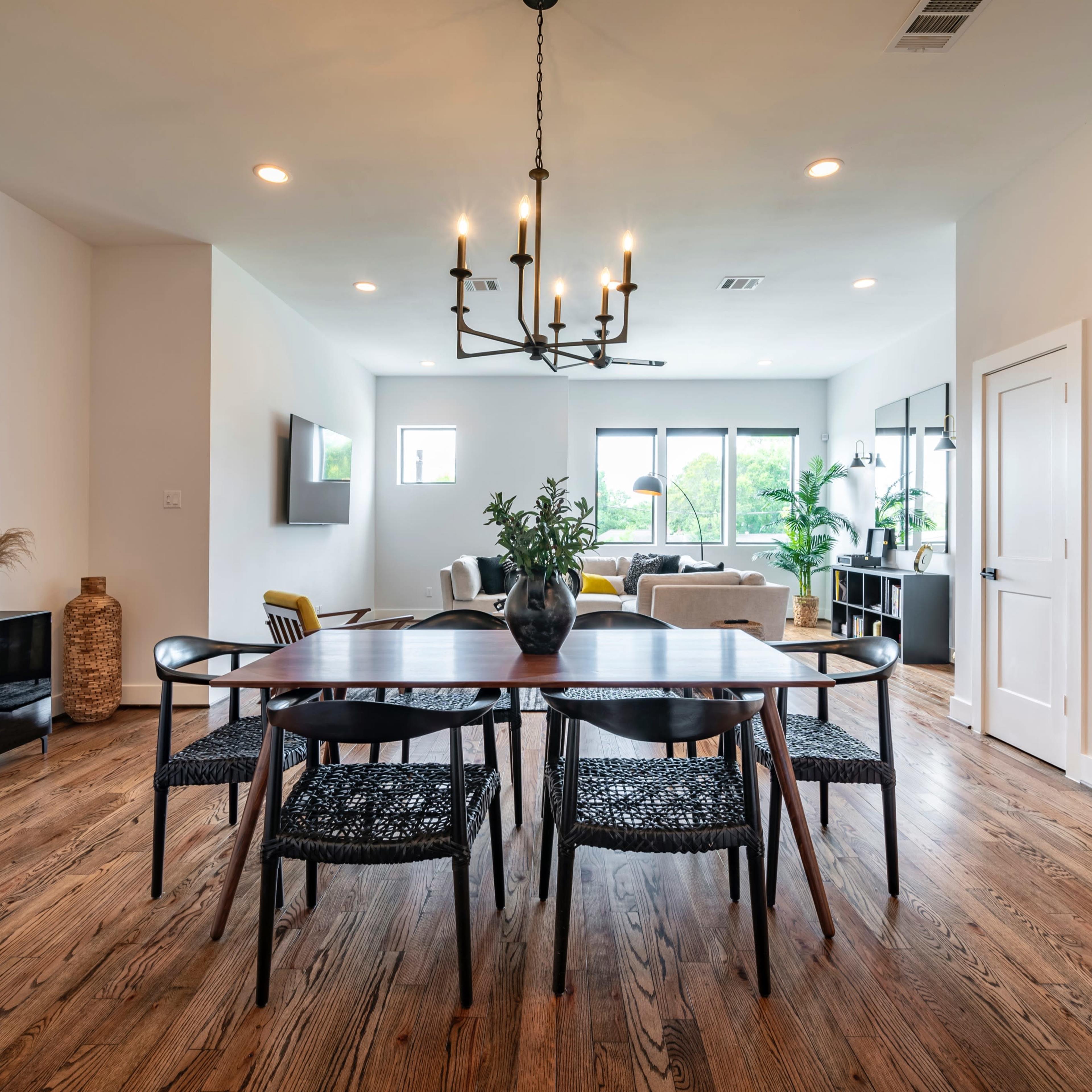 A dining area features a dark wood table surrounded by black chairs, with a modern lighting fixture above and an open layout leading to a living space.
