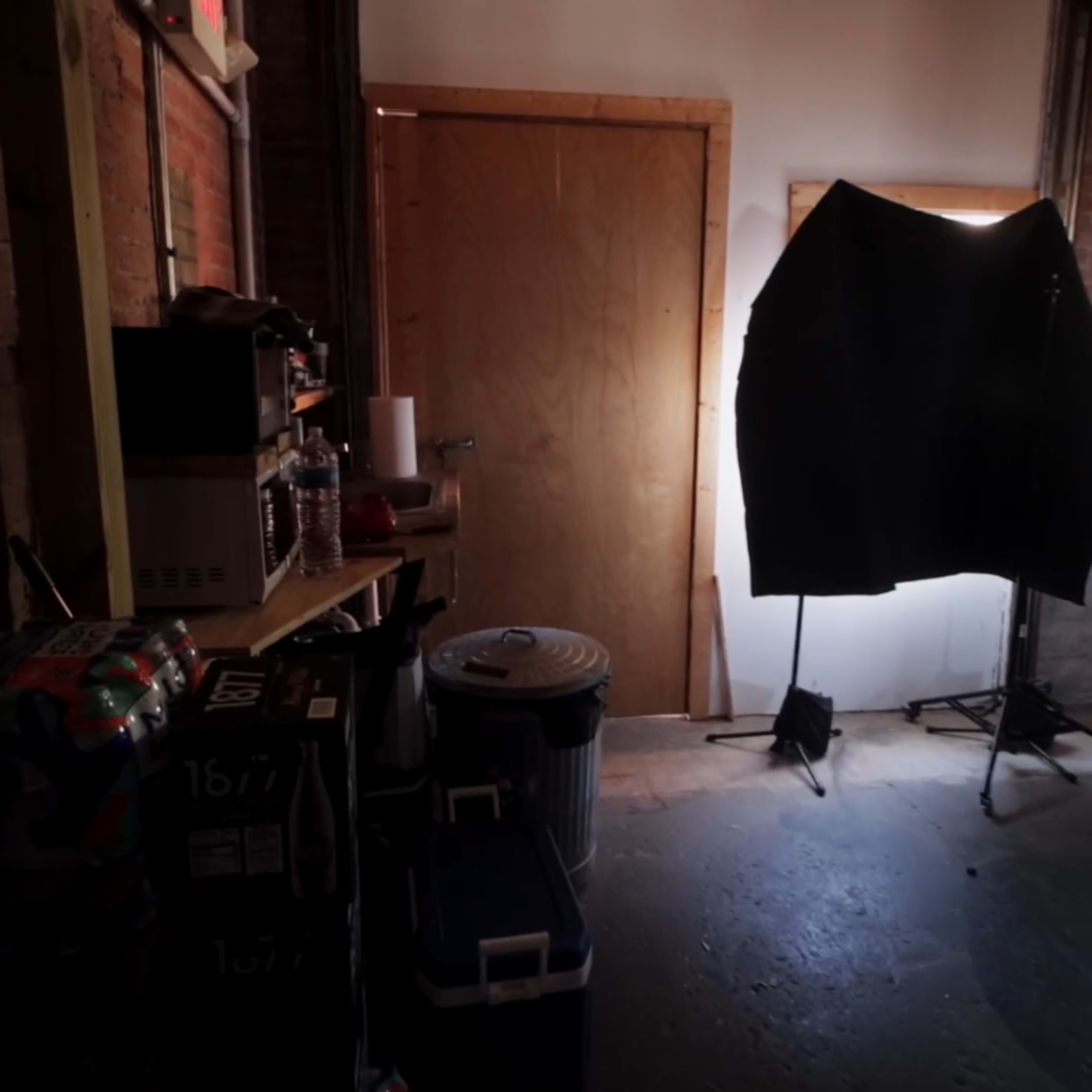A dimly lit room with wooden walls, a closed door, a black backdrop on a stand, and various equipment and containers lining a shelf.