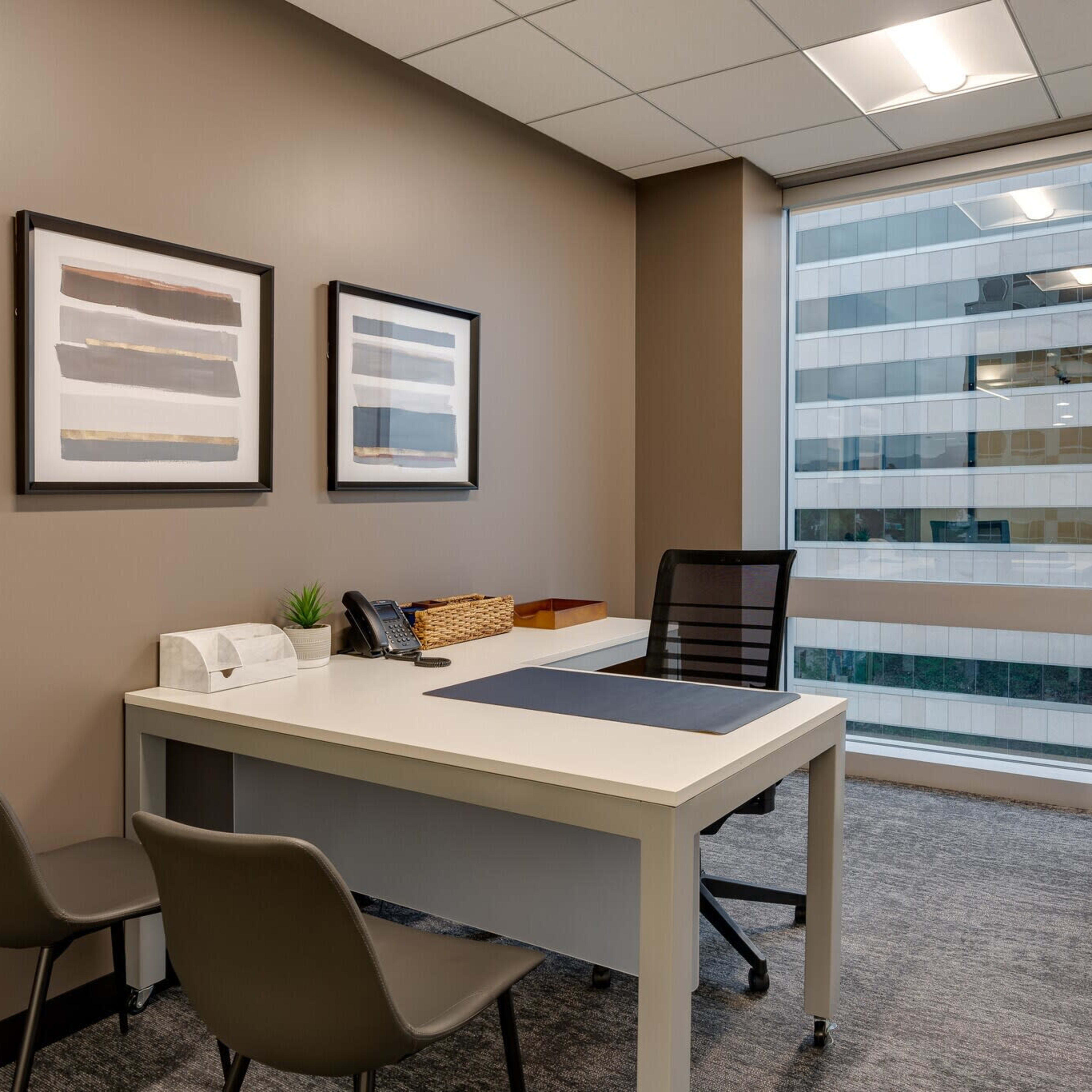 An office space featuring a desk with a chair, a telephone, and framed artwork on the walls, with a window revealing a view of a cityscape.