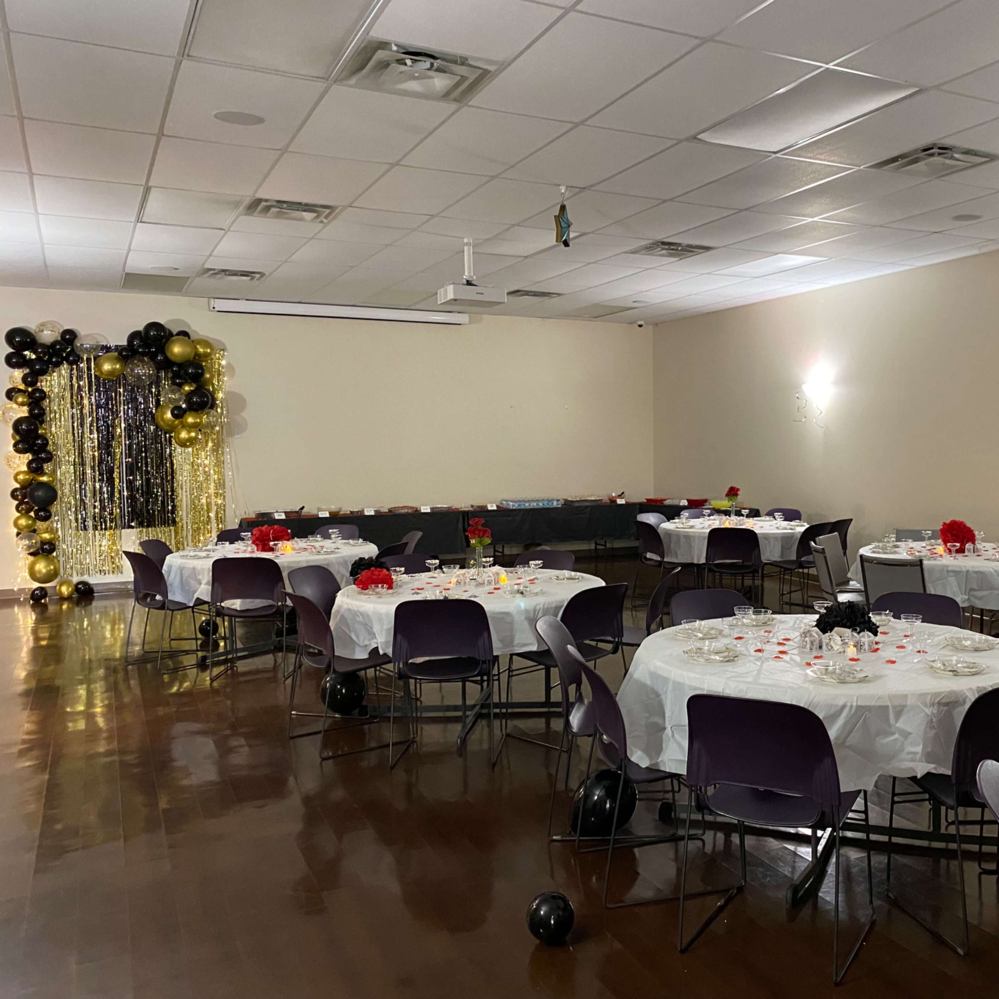 A reception hall with round tables covered in white tablecloths, decorated with red flowers and candles, and a backdrop of black and gold balloons.